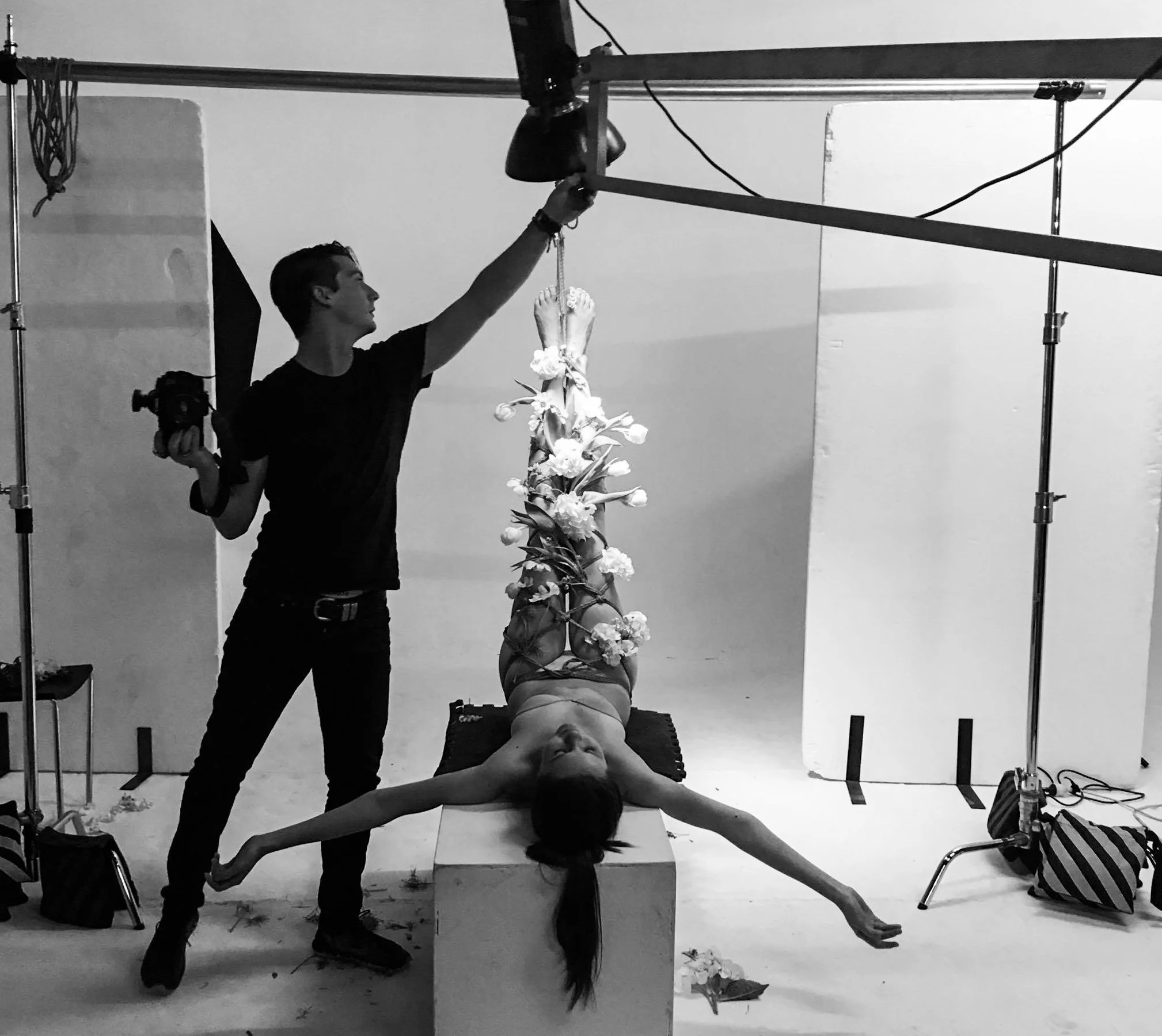 A woman lying on her back on a rectangular platform, with her arms extended outward, surrounded by flowers. A man stands next to her, adjusting a lighting fixture above her. The scene appears to be in a photography studio, with equipment, bags, and a