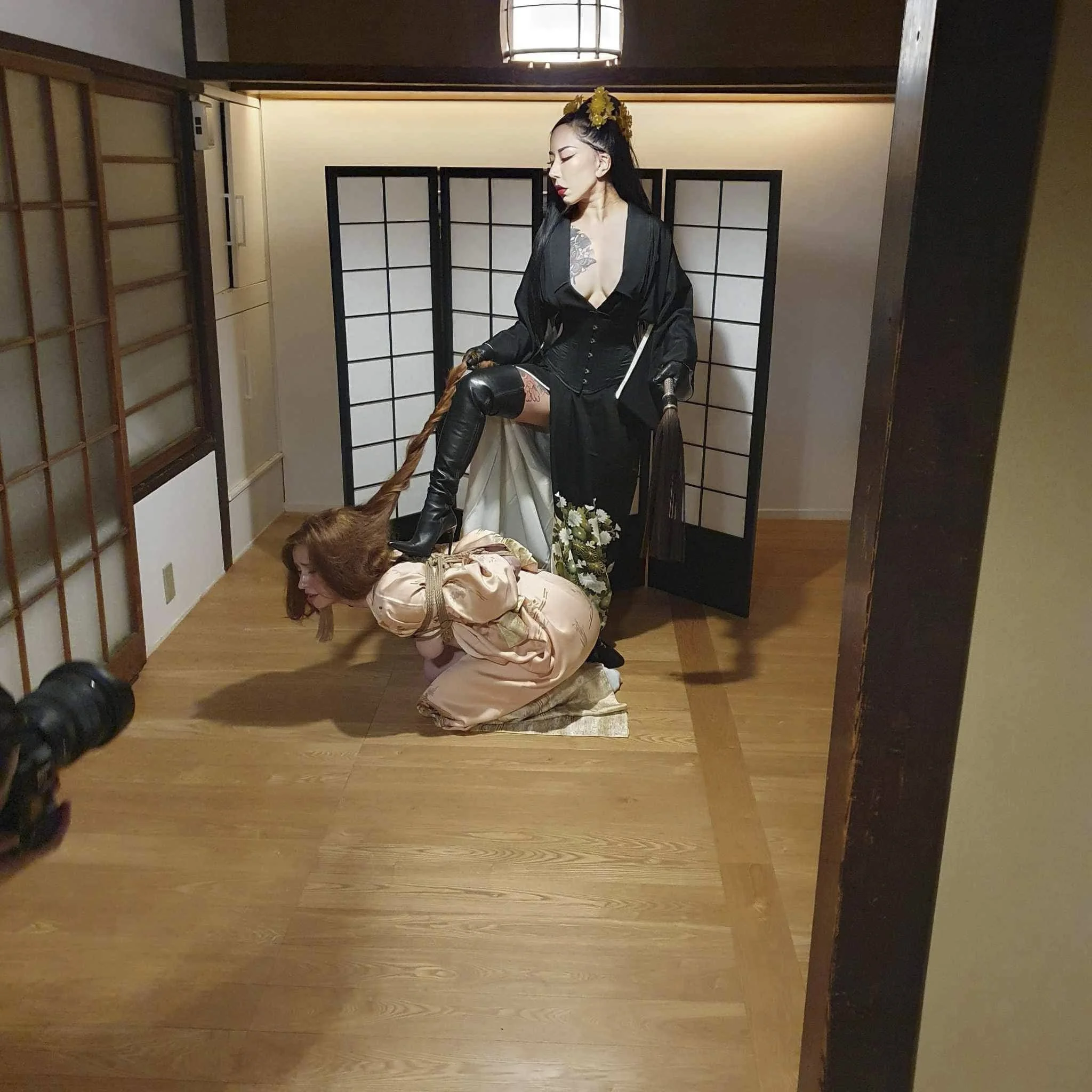 A woman dressed in black latex and a ripped black dress stands on a woman kneeling on the floor in a traditional Japanese room with shoji screens, holding a whip and a book.