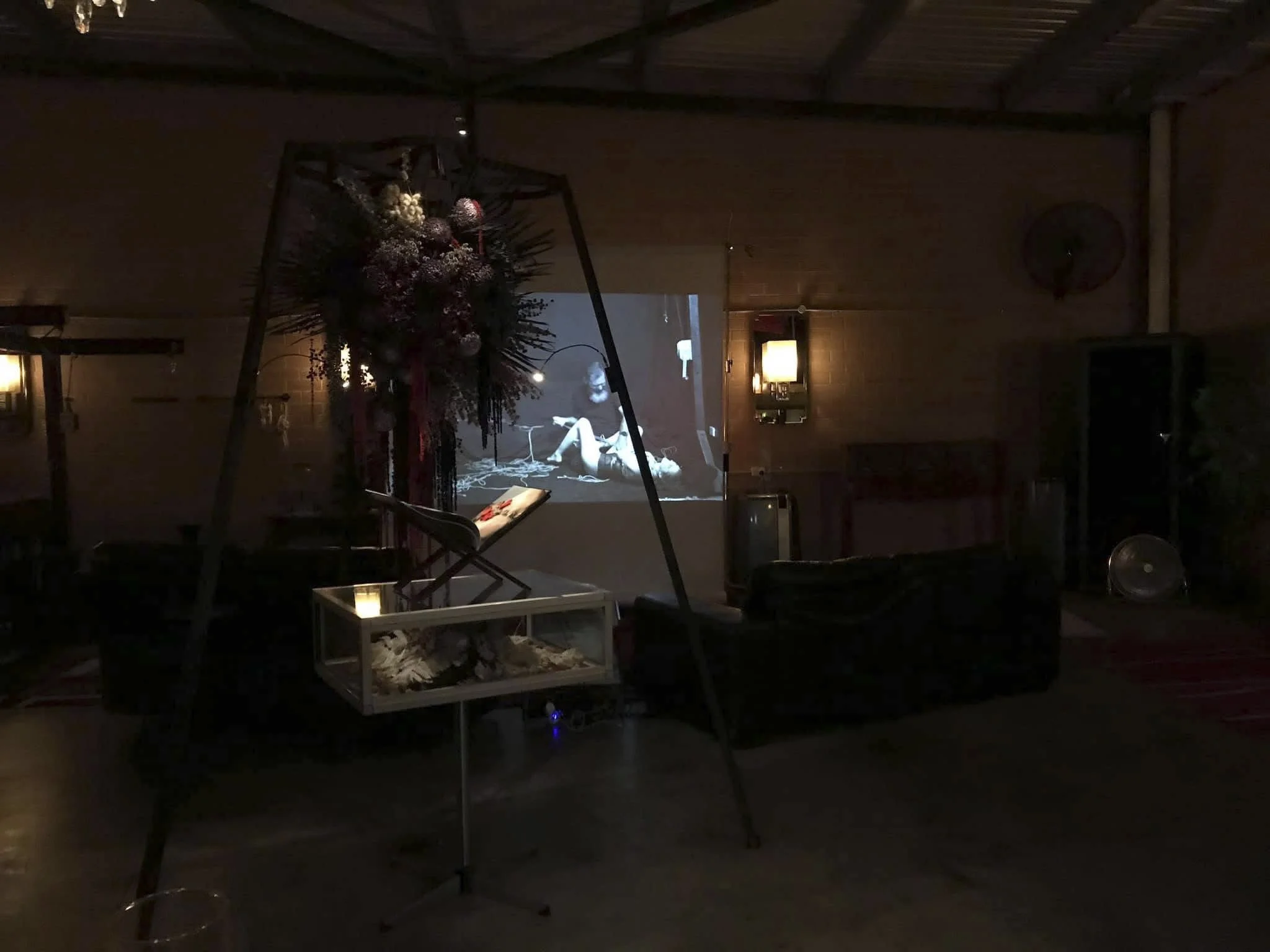 Dimly lit room with a floral decoration on a stand, a glass display case, and a projection of a person seated and playing an instrument or equipment on a wall. The room has wood-paneled walls, a sofa, and wall sconces.
