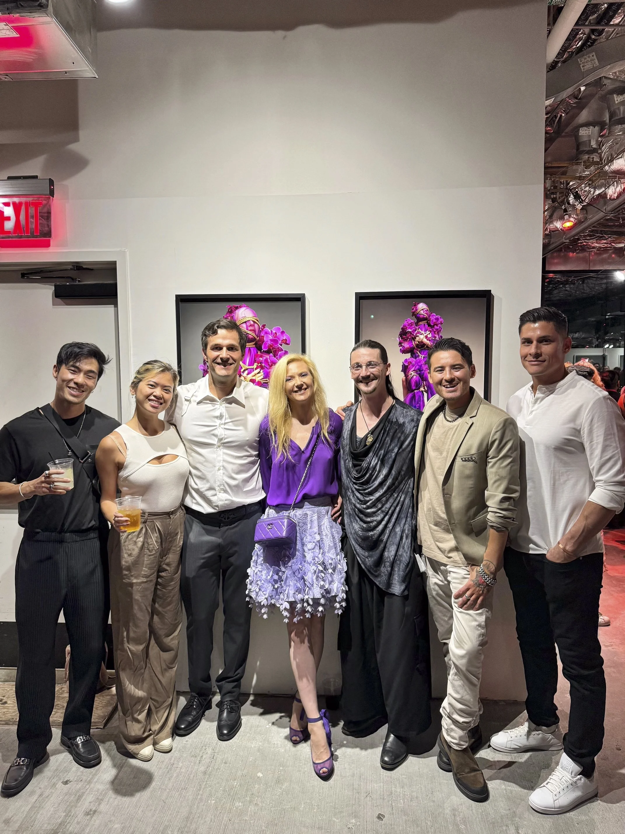 Group of eight diverse people standing together at an art gallery, smiling, with pink floral artwork in the background, some holding drinks, dressed in stylish casual and semi-formal attire.