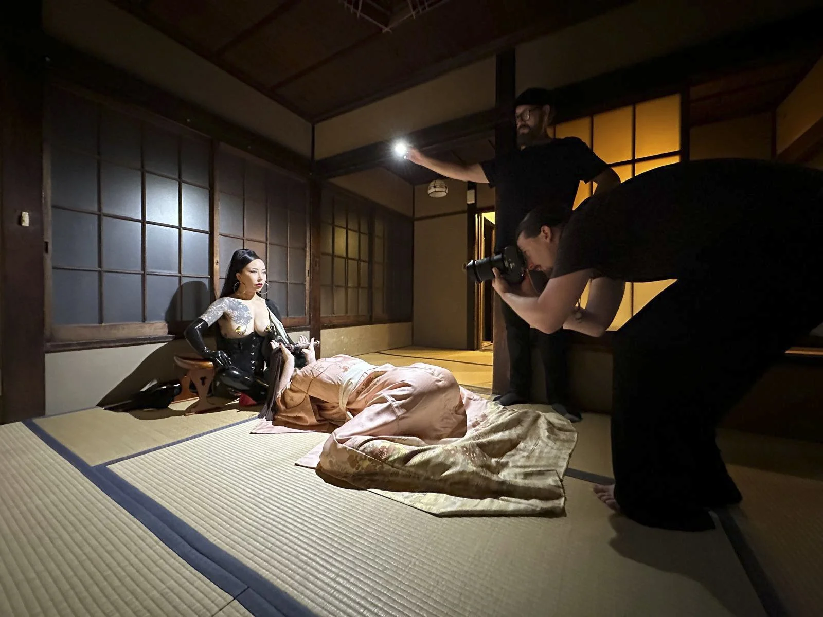 A photoshoot taking place in a traditional Japanese room with tatami mats, featuring a woman in a fancy outfit being photographed by a photographer, while an assistant holds a light and a person with a camera observes.
