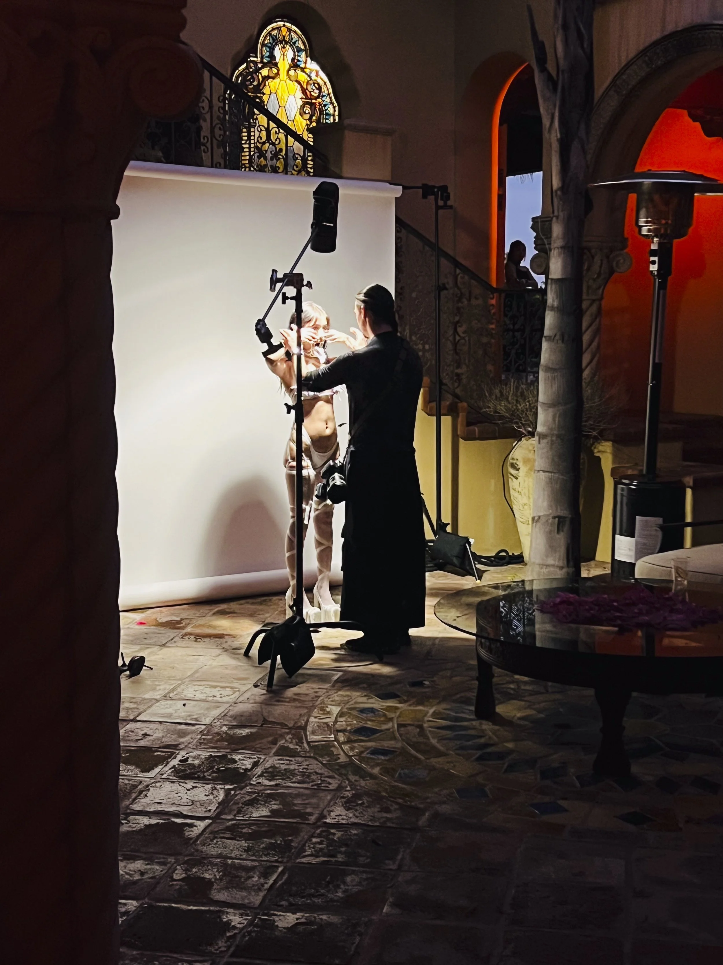 An indoor photo shows a photoshoot setup with a model and photographer by a white backdrop. The model is partially dressed, with some clothing and accessories being adjusted or applied. Studio lights and equipment are visible, and a small audience wa