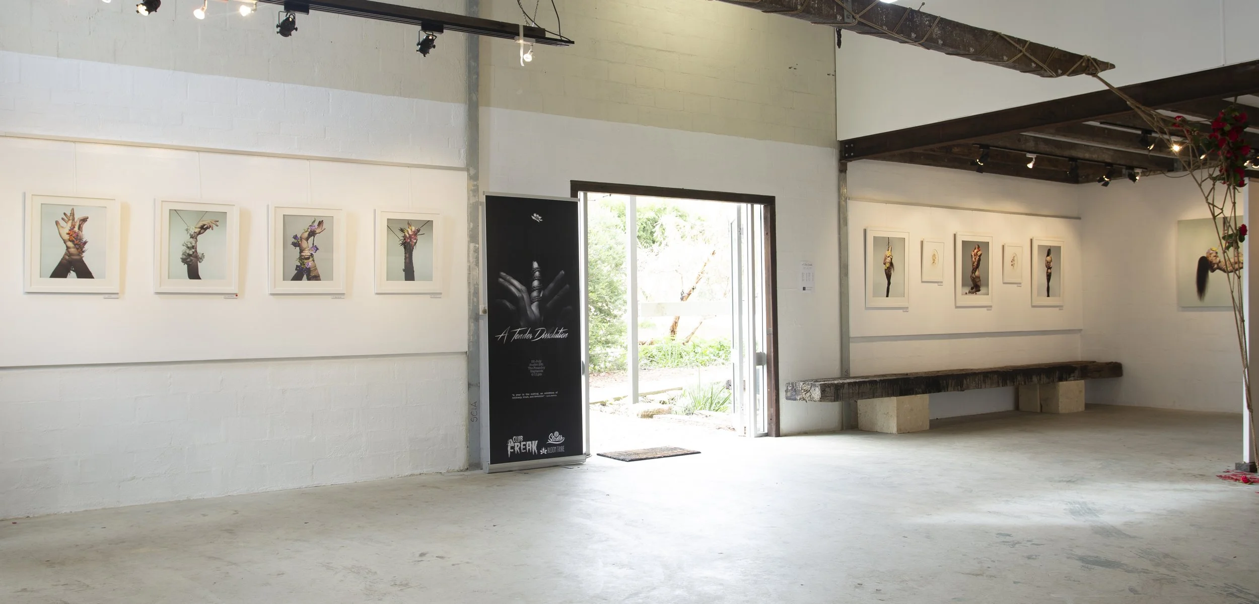 Interior of an art gallery displaying framed photographs of hands with jewelry and flowers, with a black event poster near the door and a view of greenery outside.