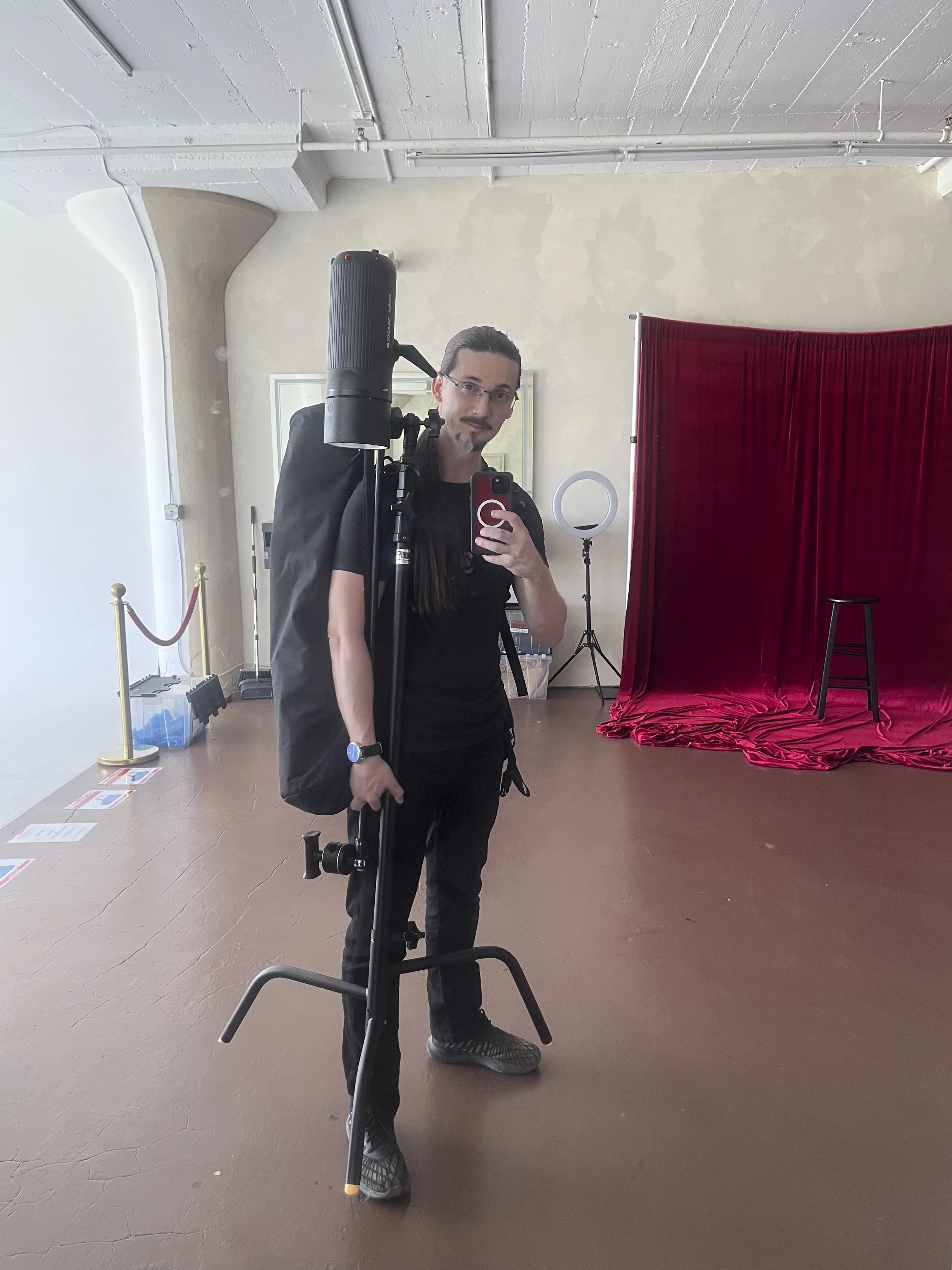A man with glasses and a mustache taking a selfie in front of a large mirror, holding photography equipment including a tripod and light. The background features red curtains, a stool, and a ring light.