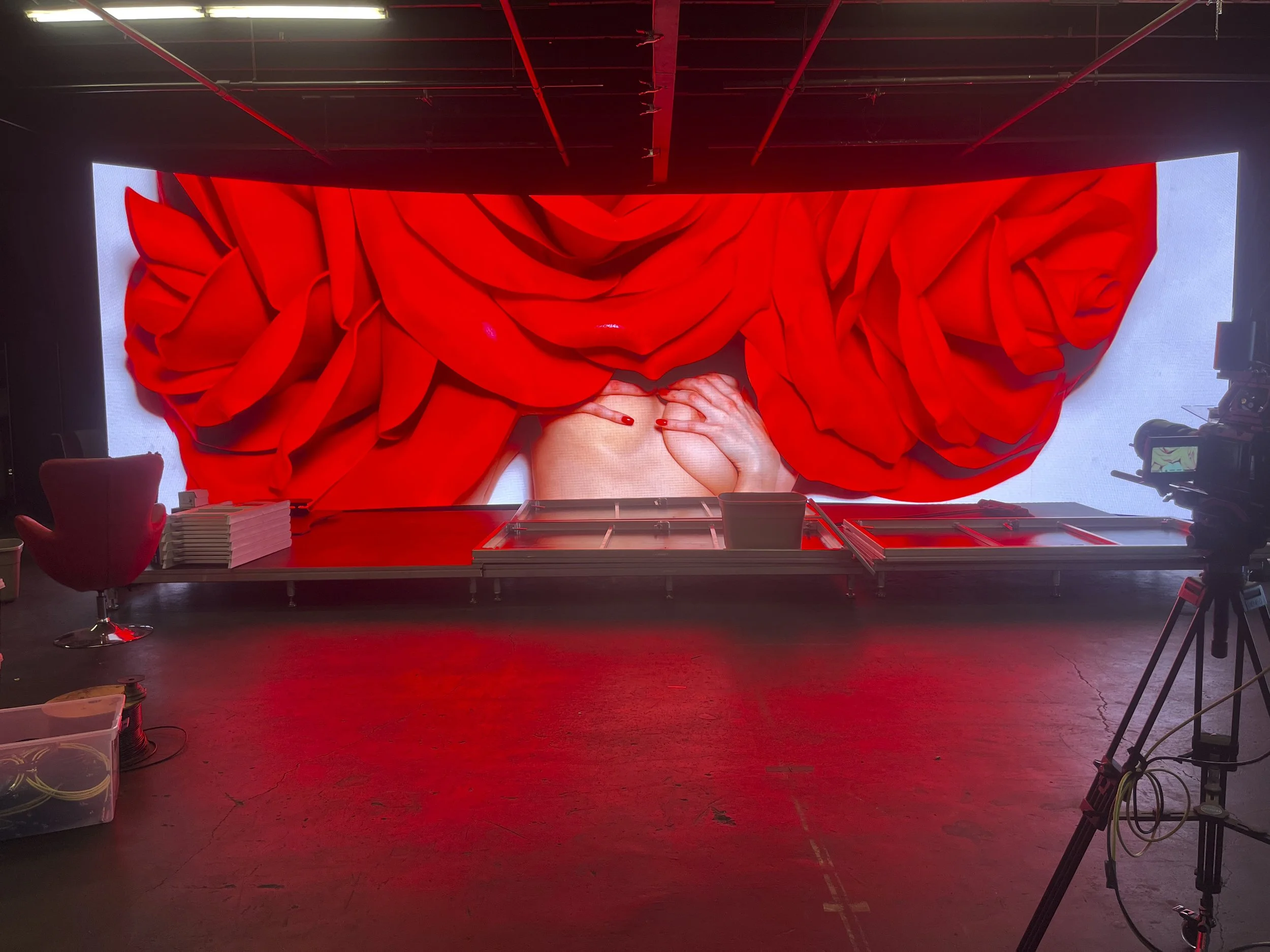 A large digital display with red fabric and hand imagery, likely part of a stage or art installation, inside a room with a camera on a tripod.