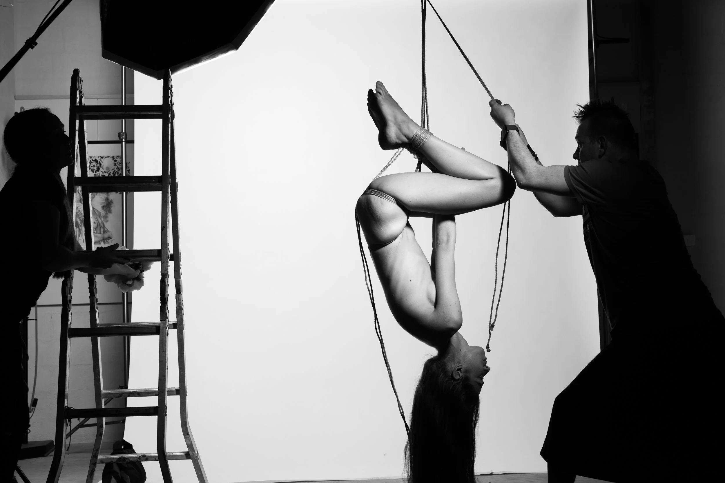 A woman's body is suspended upside down, hanging from ropes during a photoshoot, with a photographer and assistant working in a studio with lighting and a plain backdrop.