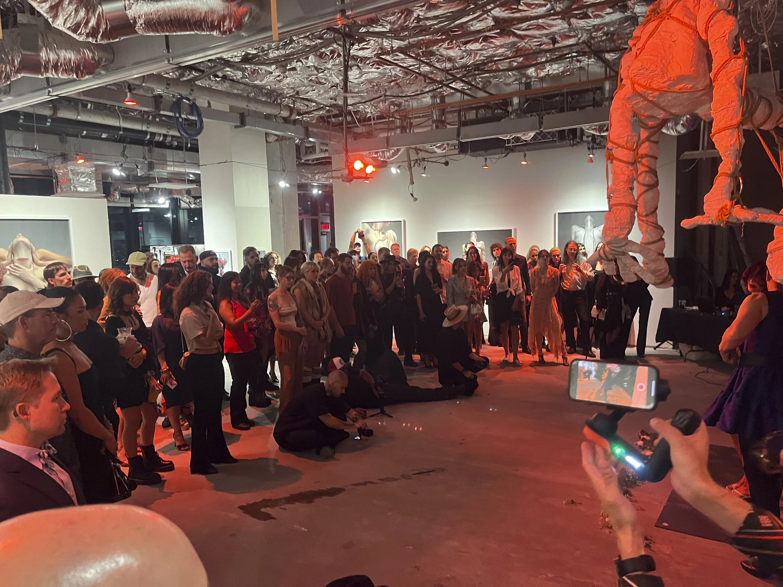 Guests attending an art gallery opening or exhibition, standing and observing artworks displayed on walls. Some people are taking photos, and the room has industrial ceilings with exposed pipes. The lighting is dim with red and white lights.