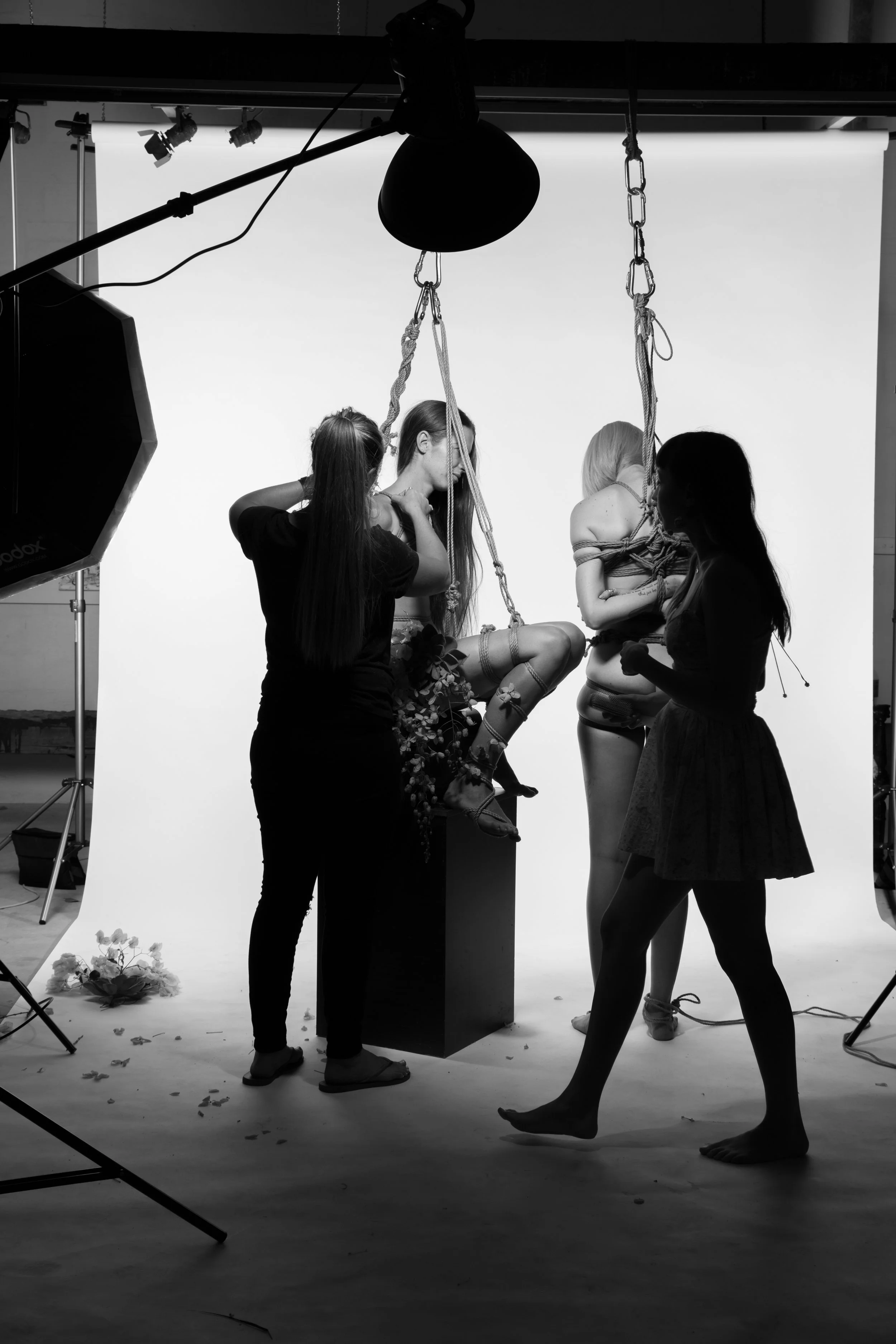 Photographers and models during a photoshoot with a white background, featuring two female models with ropes, flowers, and makeup, in a studio setting.