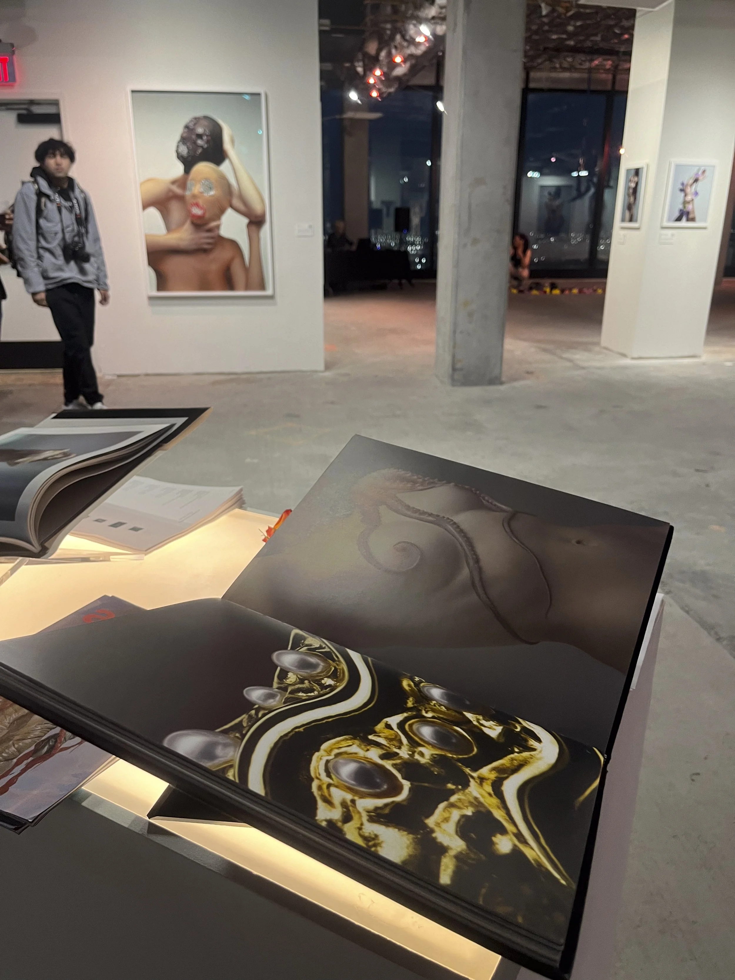 Open art book on a table displays abstract and surreal artwork, with a contemporary art gallery in the background featuring large colorful paintings and visitors.