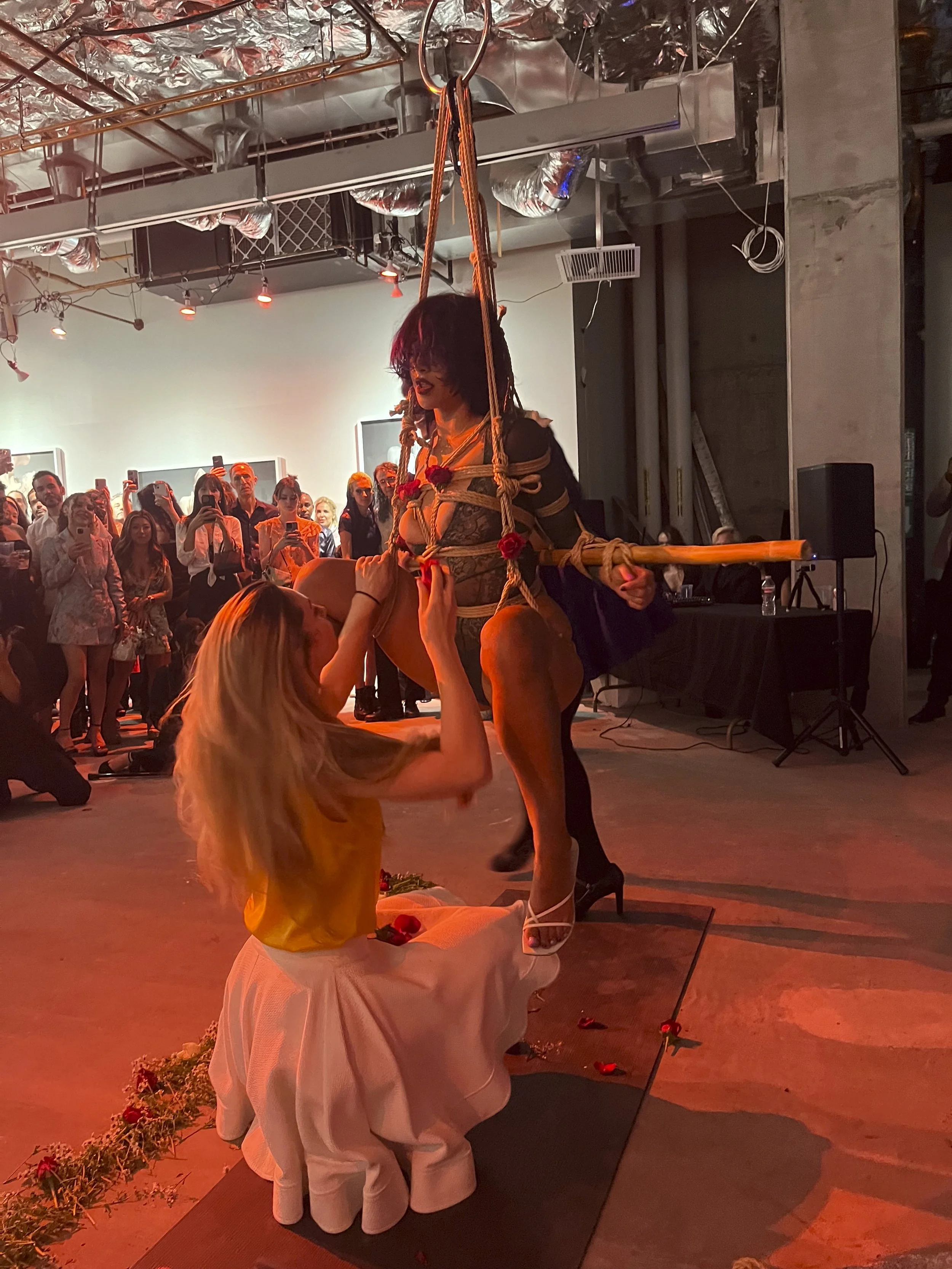 A woman in lingerie and high heels is suspended by ropes in a performance art piece, with a woman kneeling and touching her leg. An audience surrounds them, watching and taking photos in an industrial-style venue with exposed ductwork and concrete wa