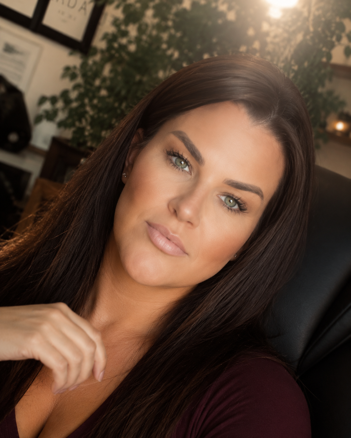 Close-up of a woman with long dark hair and blue eyes looking at the camera, indoors with plants and framed pictures in the background.