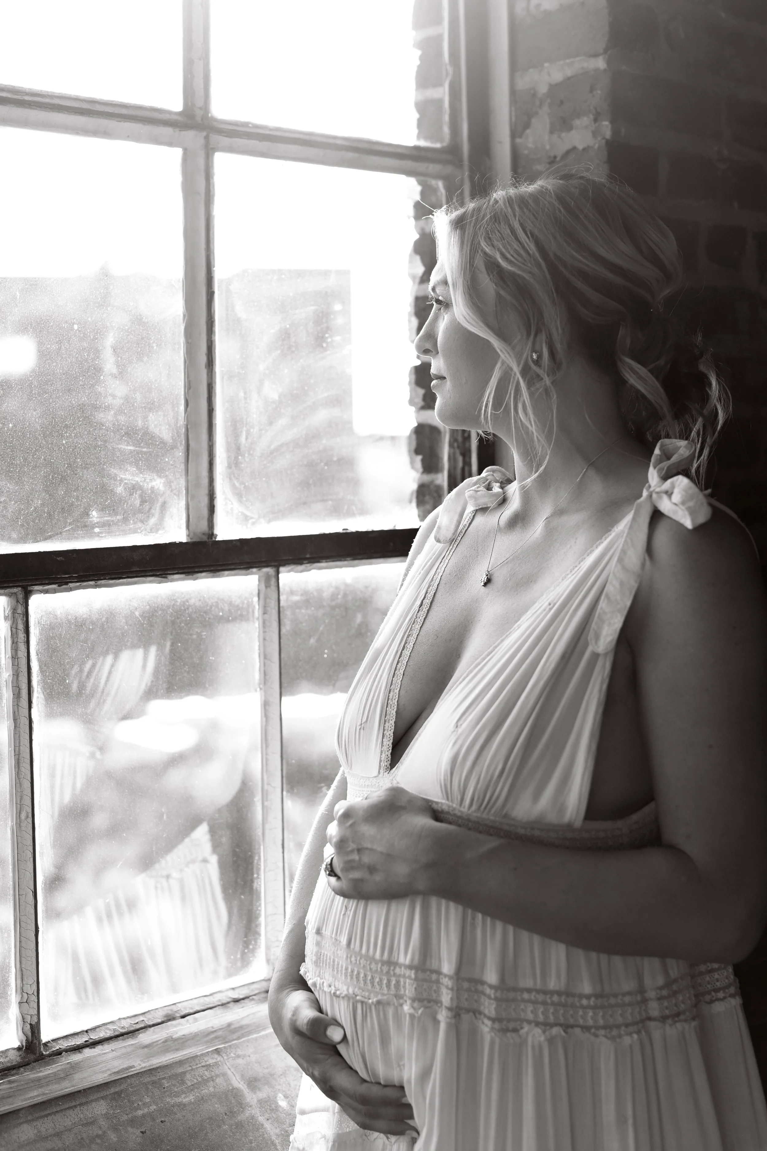 Black and white photo of a pregnant woman looking out a large window with sunlight streaming in, wearing a light-colored dress with shoulder ribbons.