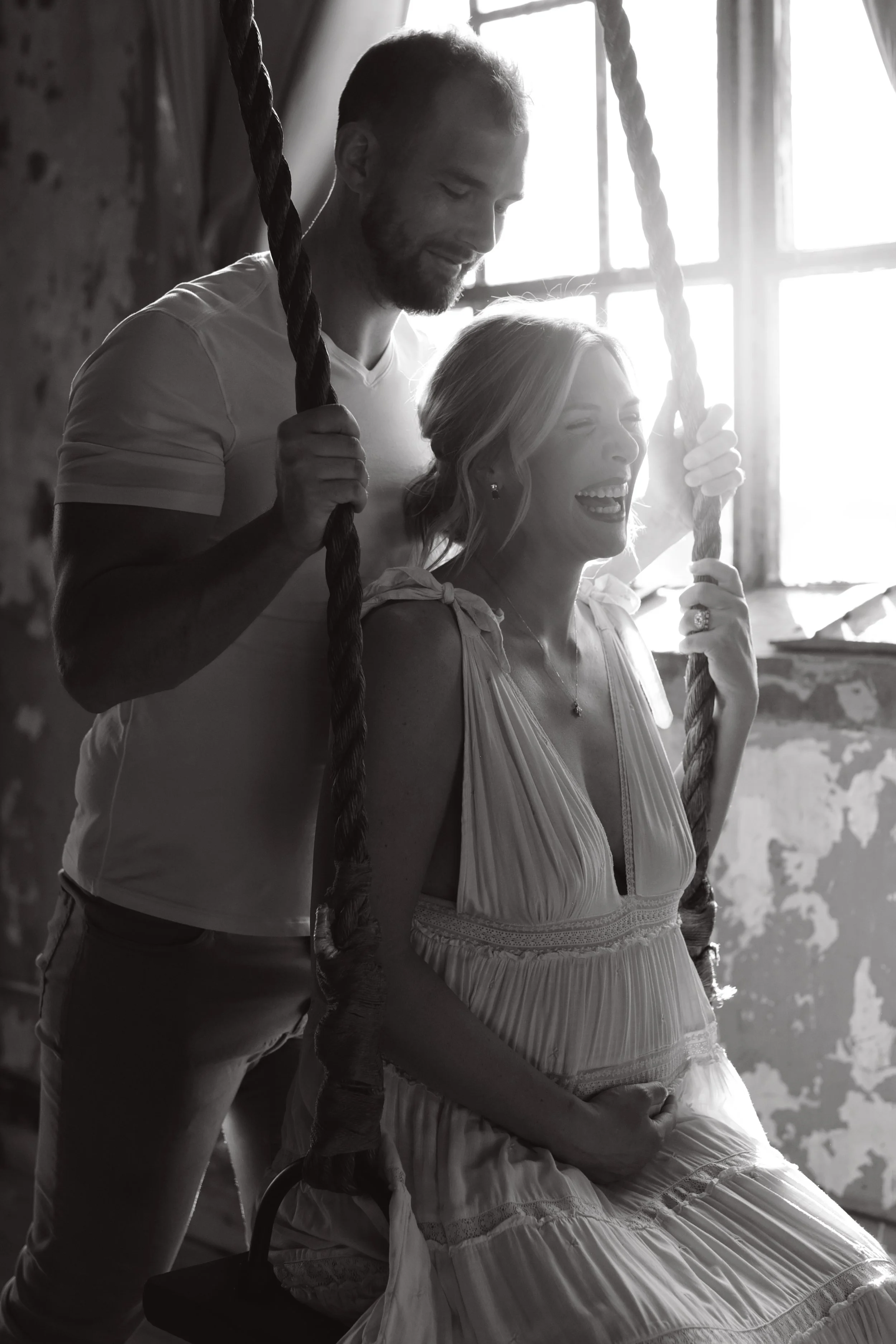 A woman sitting on a swing held by a man inside a sunlit room with large windows and worn walls, both smiling.