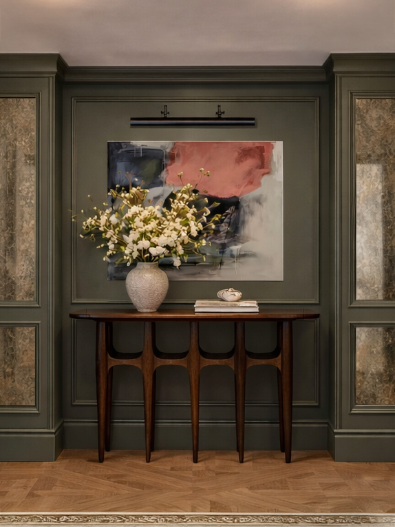 Interior wall decor featuring a green paneled wall with a large abstract painting, a wooden console table, a vase with white flowers, and a stack of books.