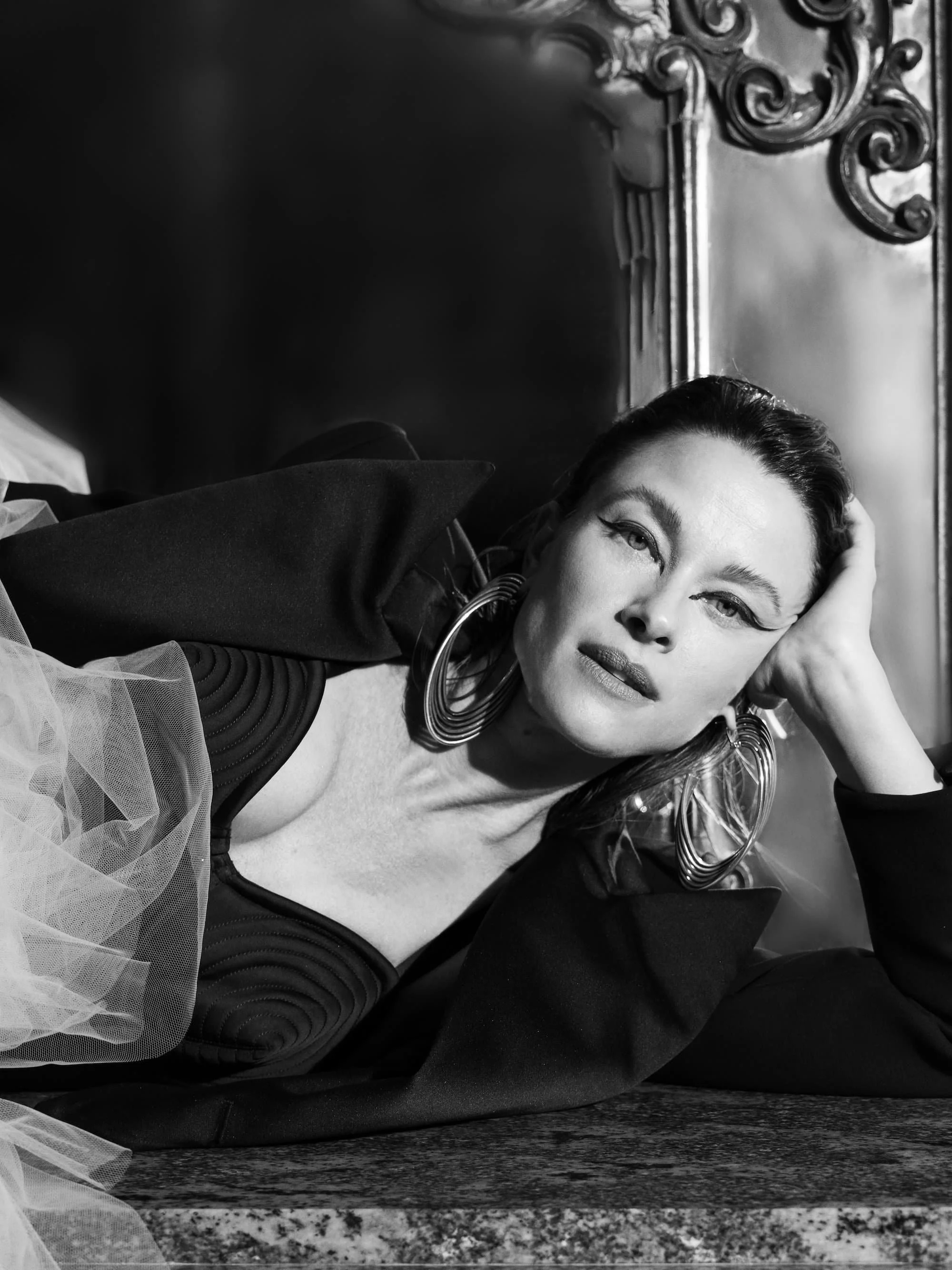 A woman with makeup and large circular earrings lying on her side on a table, resting her head on her hand, in black and white.