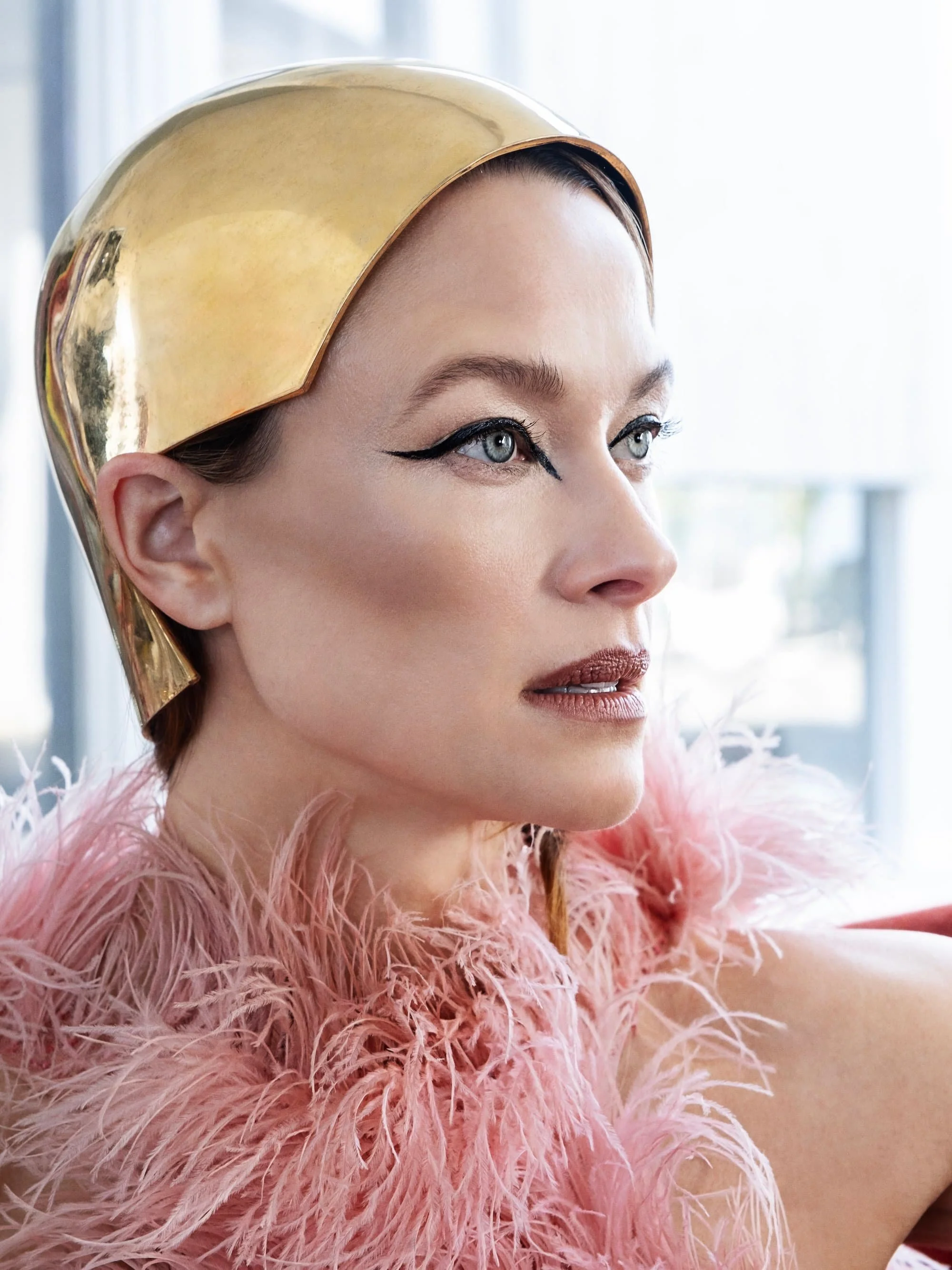 A woman with platinum blonde hair wearing a shiny gold headpiece and a pink feathered garment, looking to the side with makeup including winged eyeliner and brown lipstick.