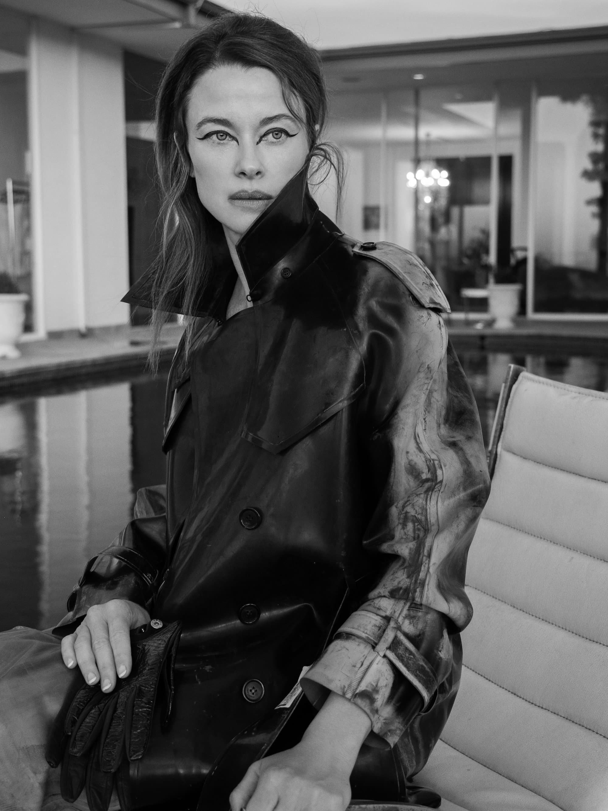 A woman with long hair and bold eyeliner sitting indoors, wearing a shiny black leather jacket with the collar up.