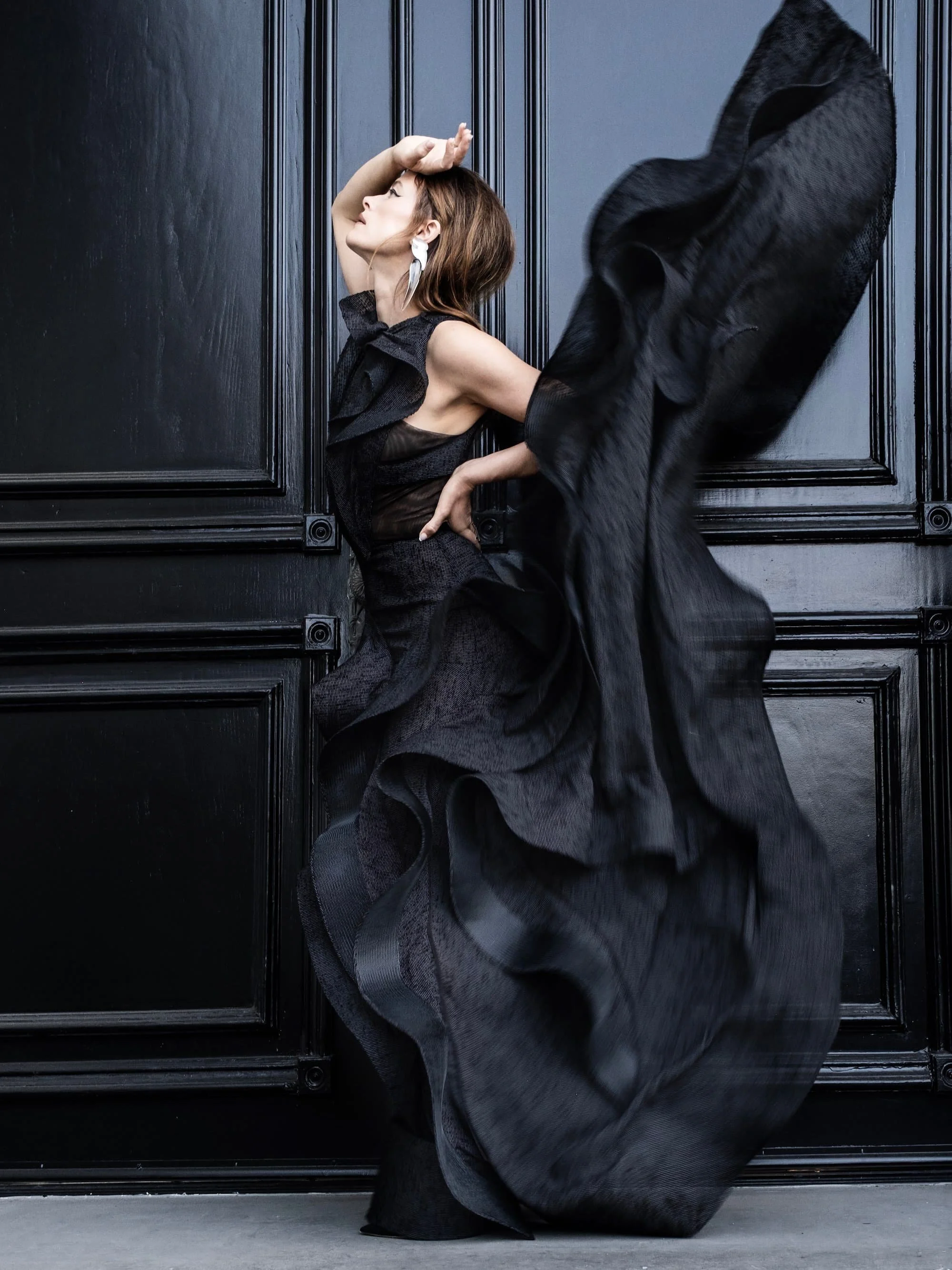 A woman wearing a black flowing dress, leaning against a black paneled wall, with one hand on her hip and the other hand touching her forehead.