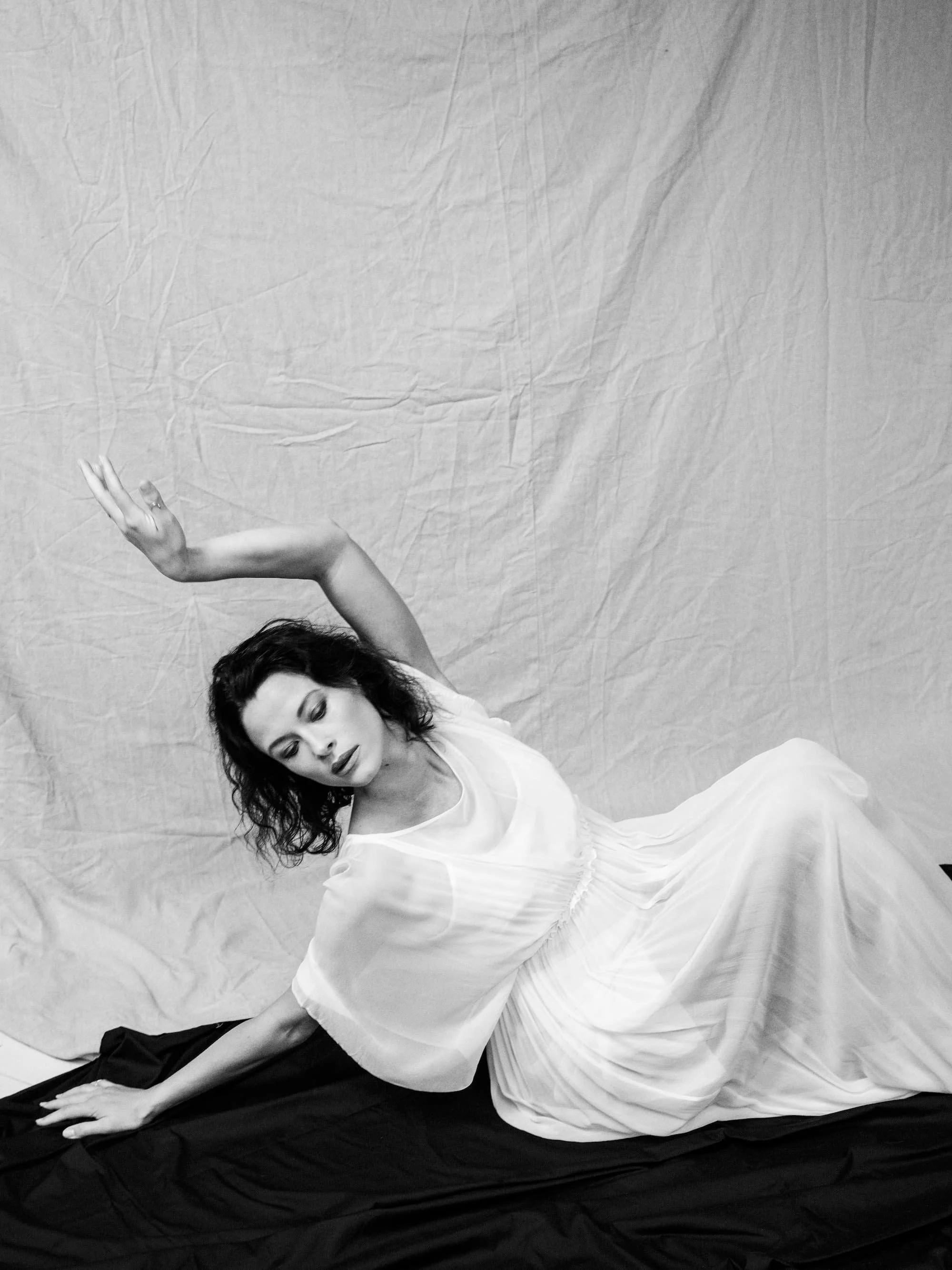 A woman in a white dress sitting on a black fabric, with her eyes closed and one arm raised above her head, against a plain backdrop.
