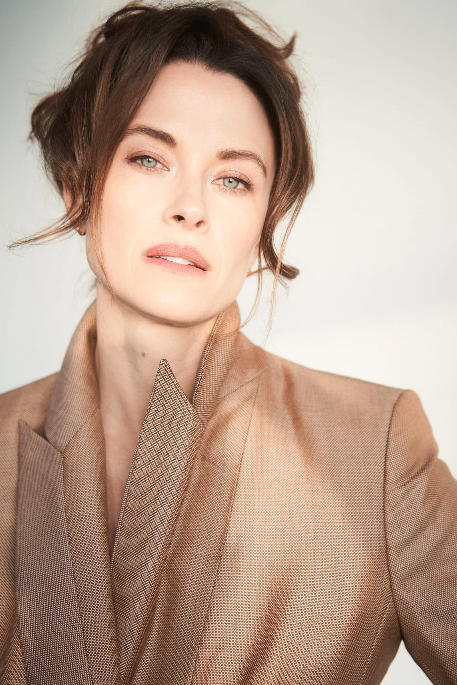 Woman with light eyes and brown hair in a tan blazer, looking at the camera with a soft expression, against a neutral background.