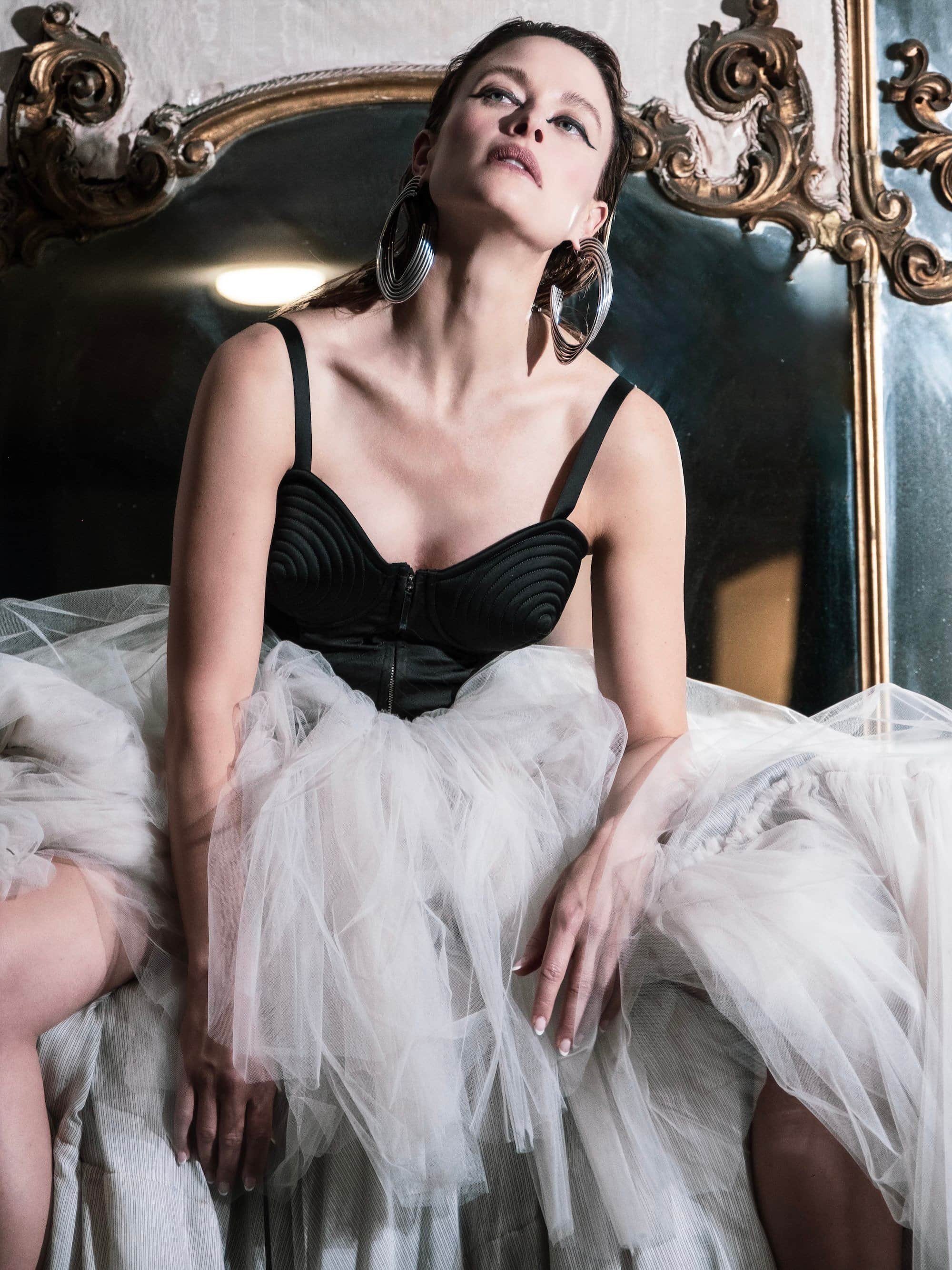 Woman sitting on a bed with a white tulle skirt, wearing a black top with spaghetti straps and large silver hoop earrings, in front of a vintage mirror.