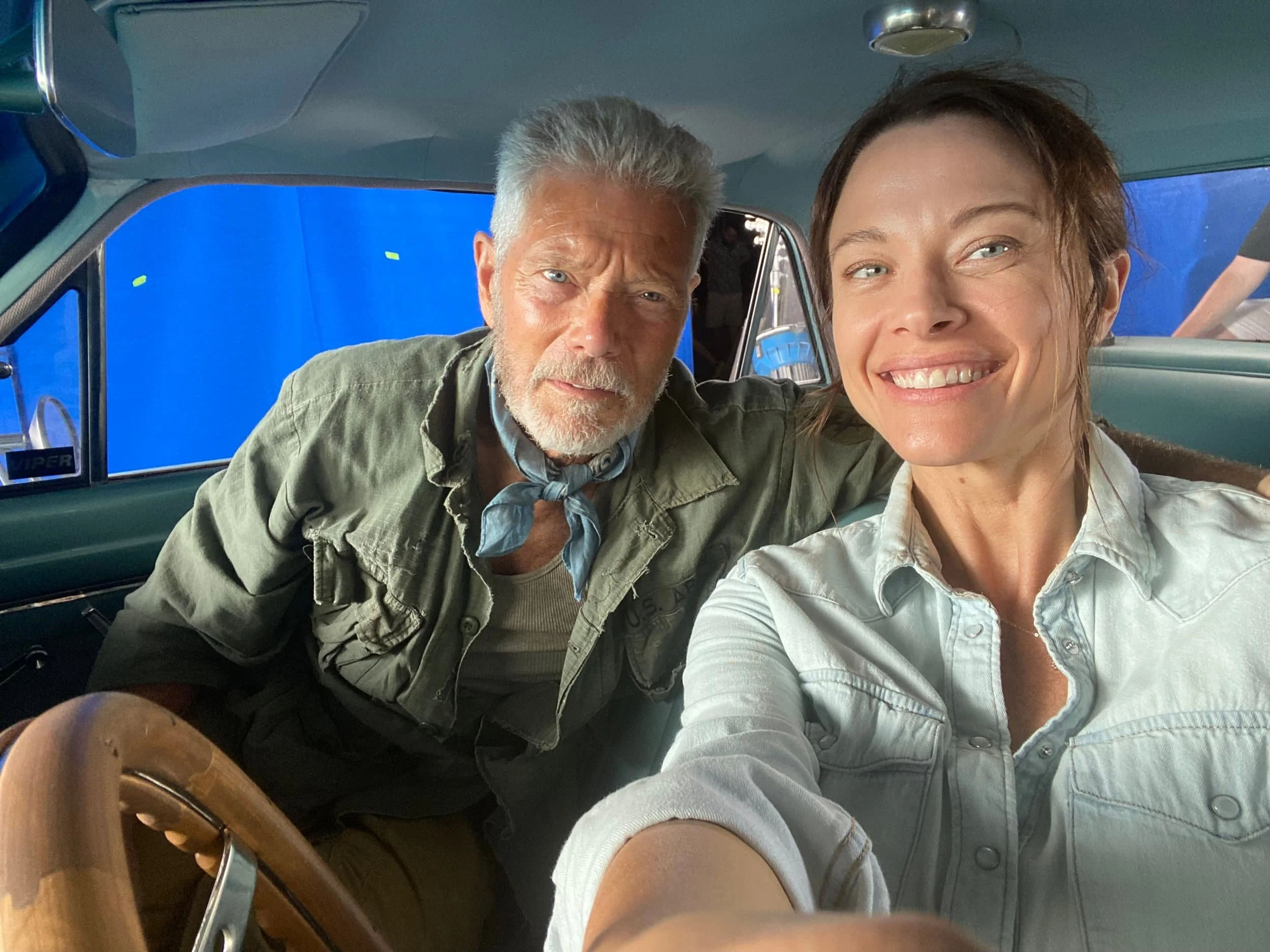 A woman and an older man taking a selfie inside a vehicle with a blue screen in the background. The woman smiles while the man appears serious.