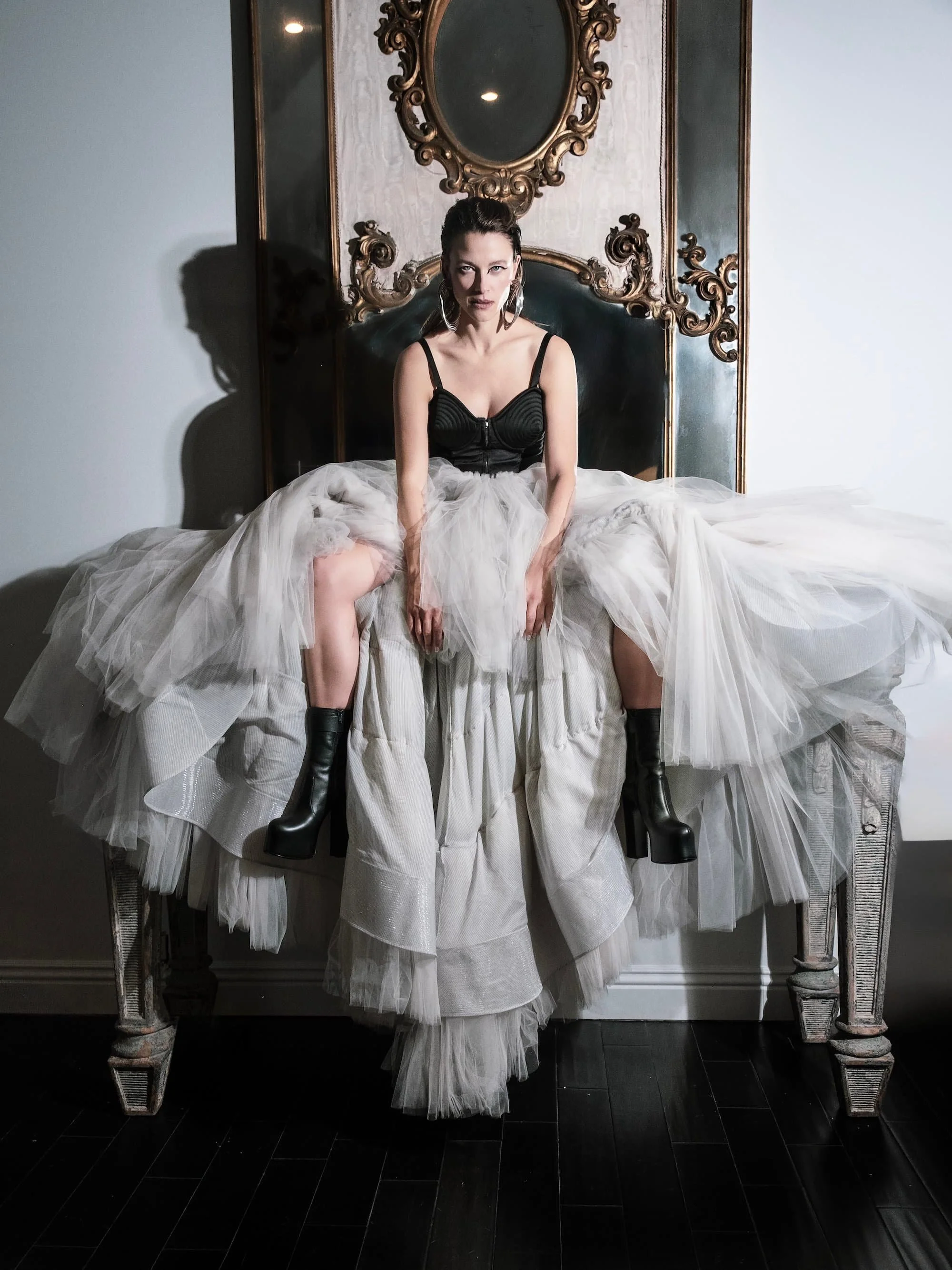 A woman sitting on a bed with a vintage frame headboard, wearing a black top, a white tulle skirt, and black boots, in a room with a mirror and decorative wall paneling.
