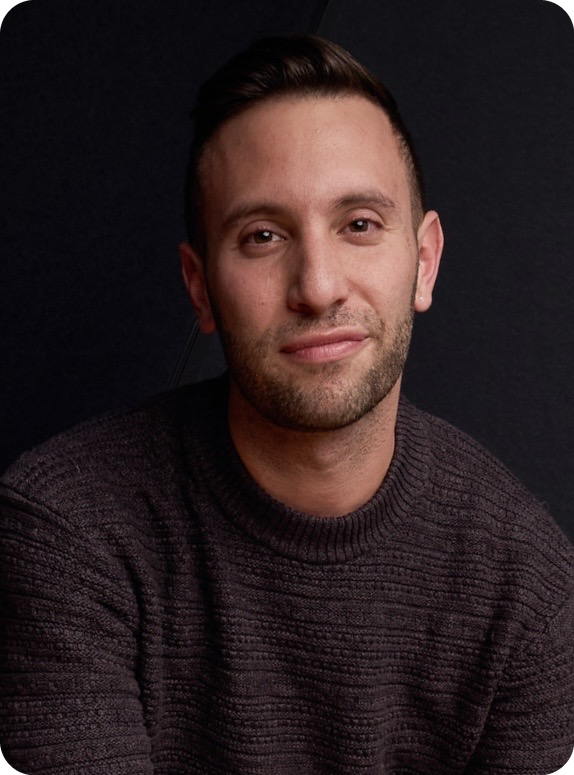 A young man with short dark hair and a beard, wearing a dark knit sweater, looking at the camera with a slight smile, against a dark background.