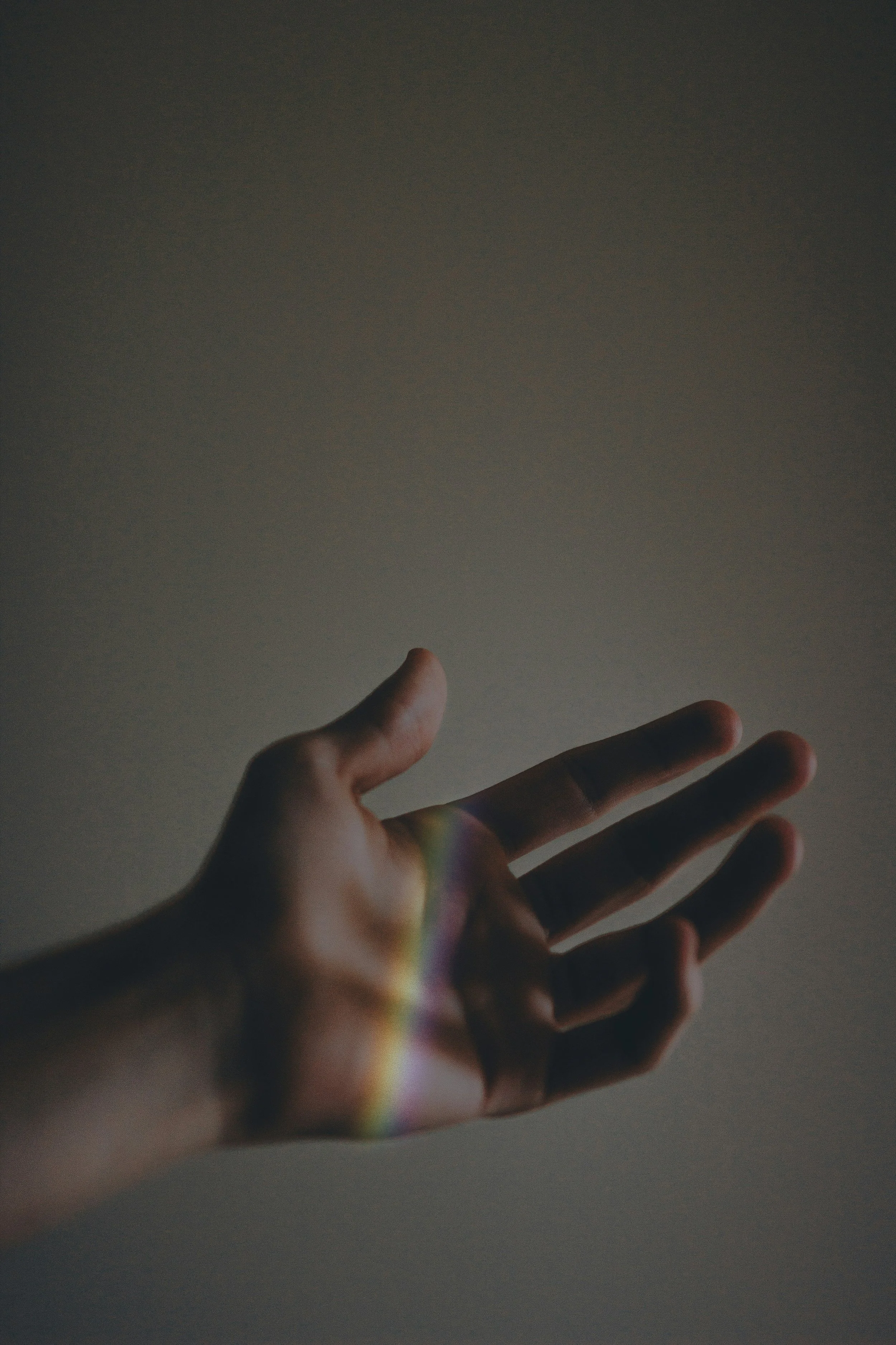 A hand reaching upward with a rainbow-colored light cast on the fingers against a gray background.