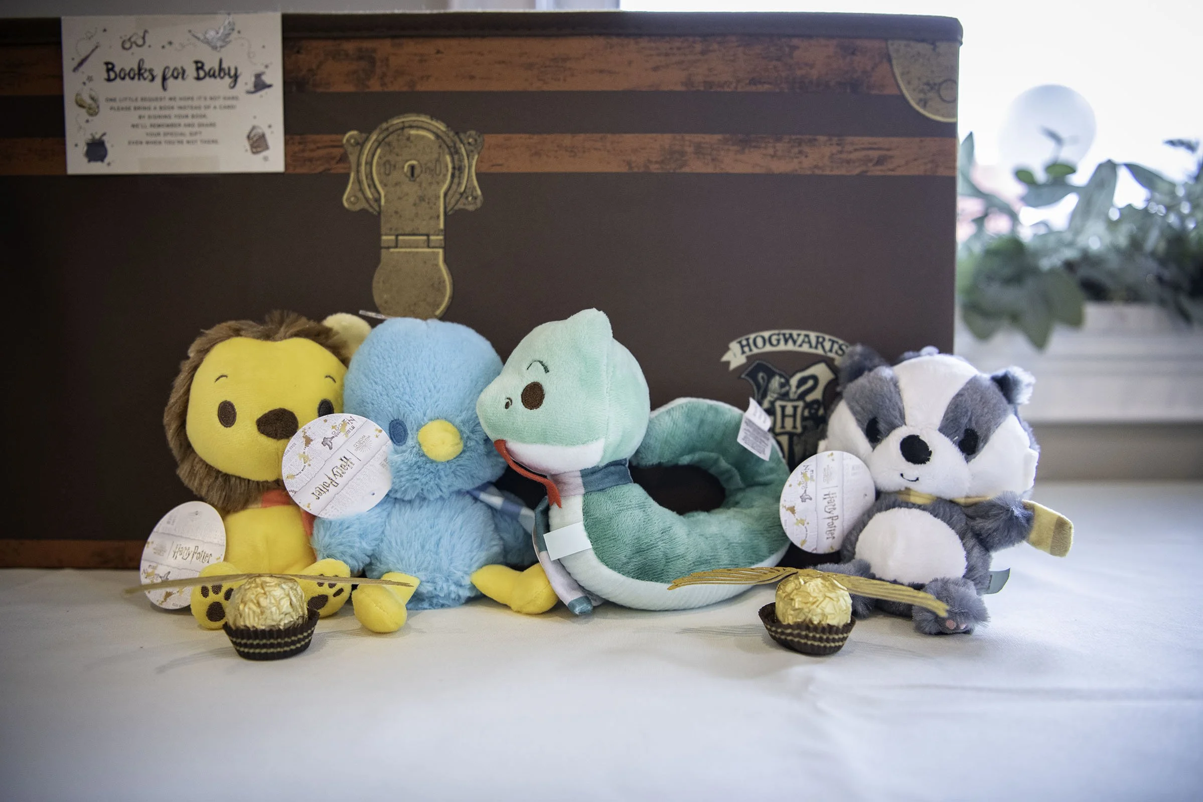 Four plush toys of characters from Harry Potter, including a lion, a blue creature with a yellow beak, a green snake with a red tongue, and a gray and white badger, arranged on a table with small chocolates and Harry Potter-themed tags in front of a 