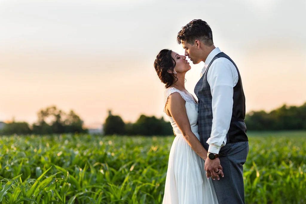 A couple dressed in wedding attire standing close in a field at sunset, with the man holding the woman's hands and their foreheads almost touching.