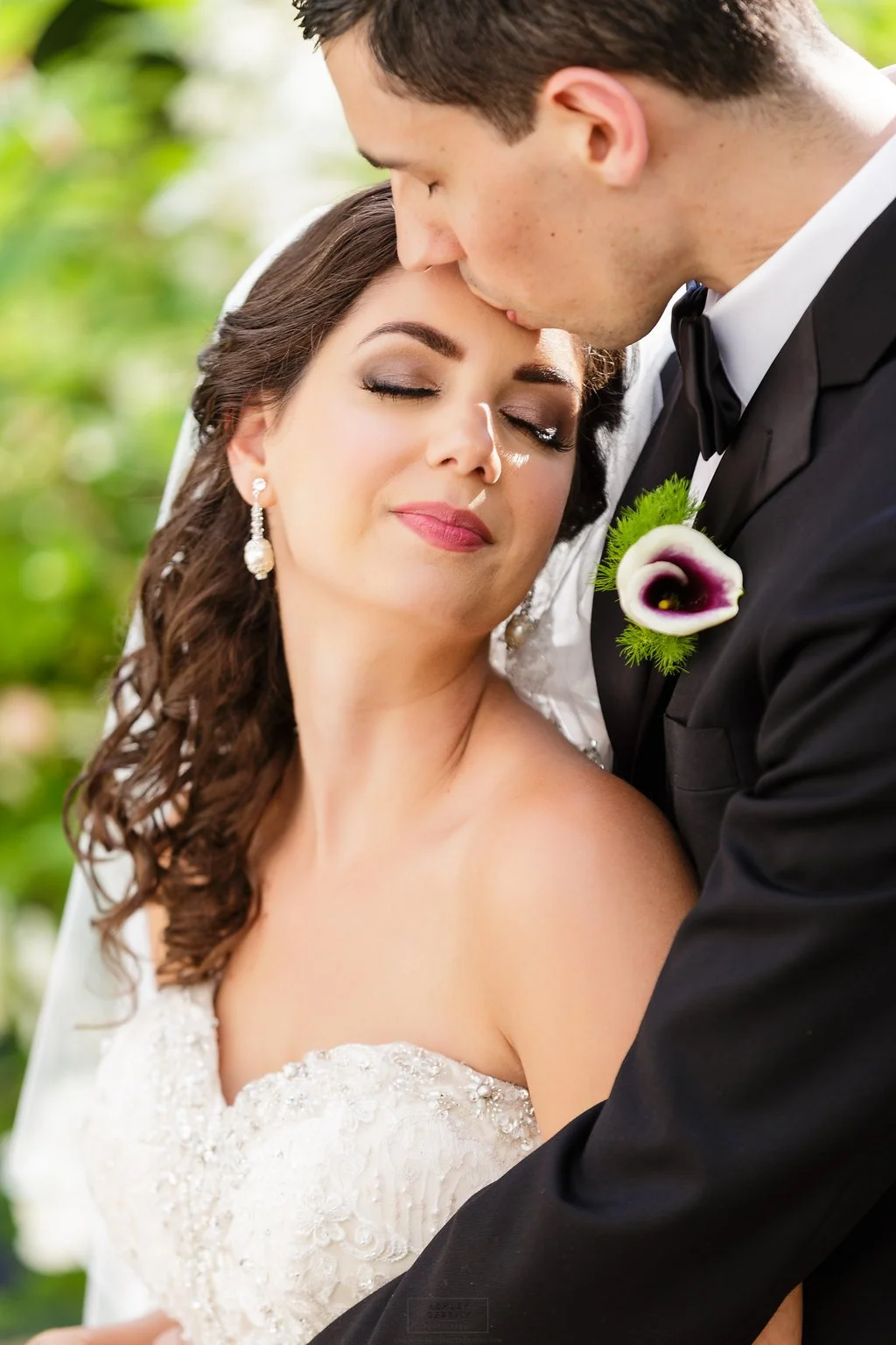 A bride and groom share an intimate moment, with the groom kissing the bride's forehead. The bride has dark, curly hair, elegant makeup, and earrings, wearing a white wedding dress. The groom is in a black tuxedo with a white shirt and bow tie, with 