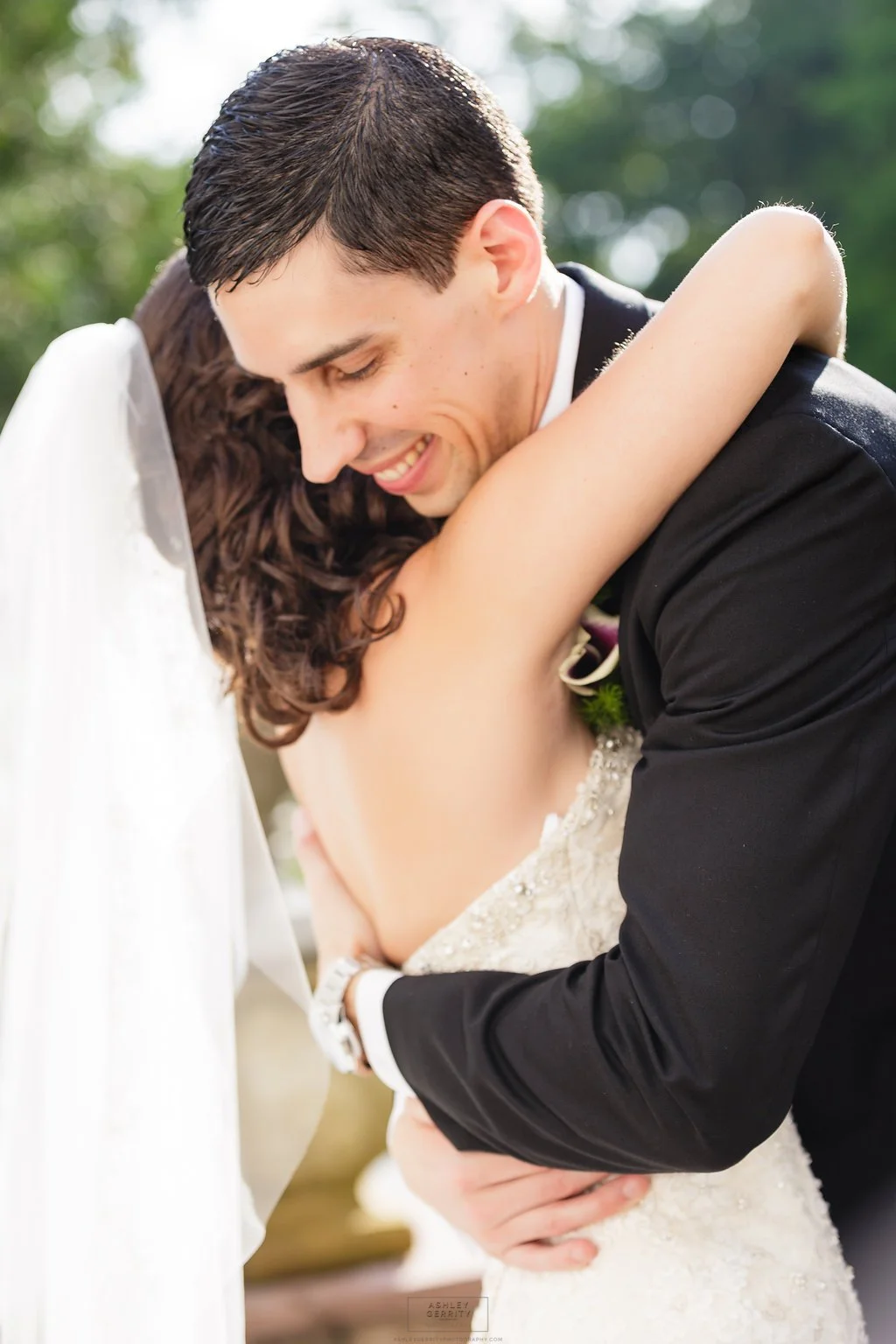 A groom wearing a black suit hugging and smiling with a bride in a wedding dress and veil.