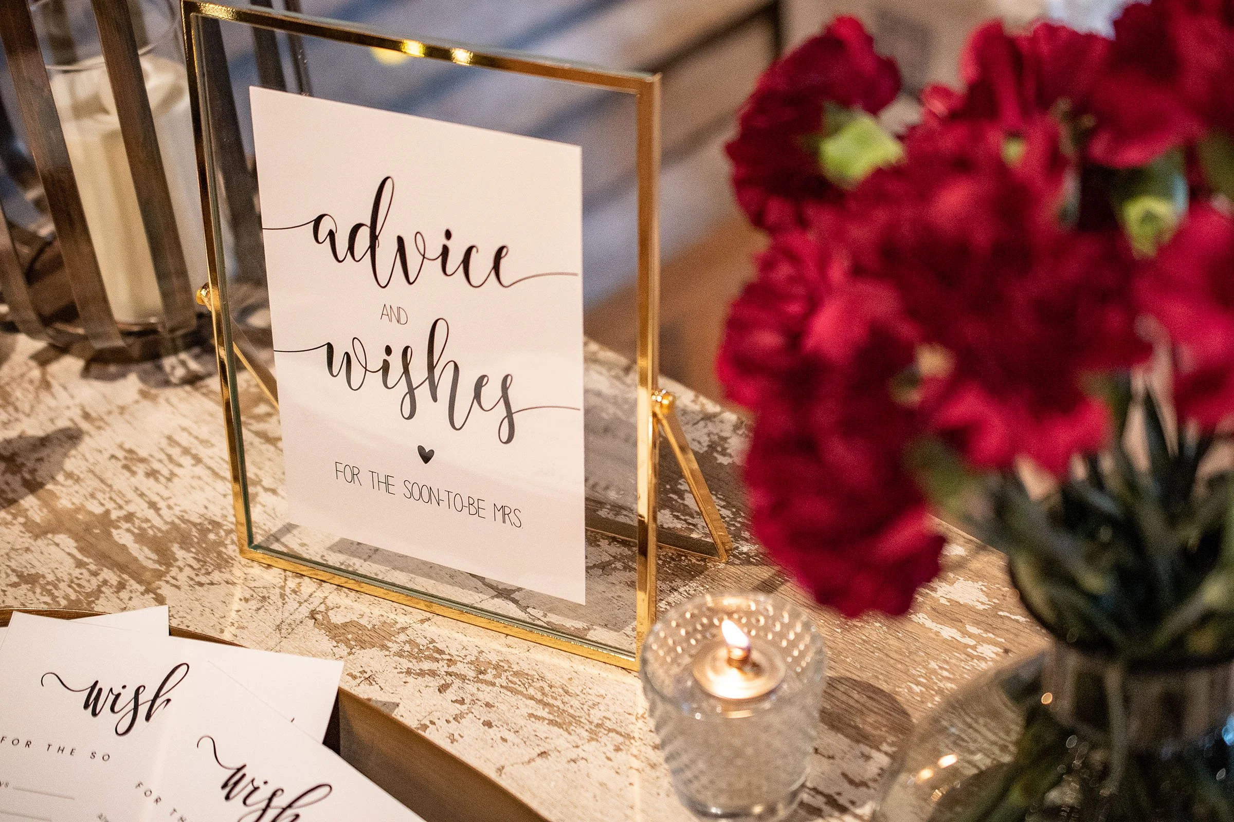 A framed sign on a rustic wooden table reads, 'advice and wishes for the soon-to-be Mrs.' with a small heart symbol beneath the text. To the right, a bouquet of deep red flowers is visible, and in the foreground, a lit candle in a glass holder adds a