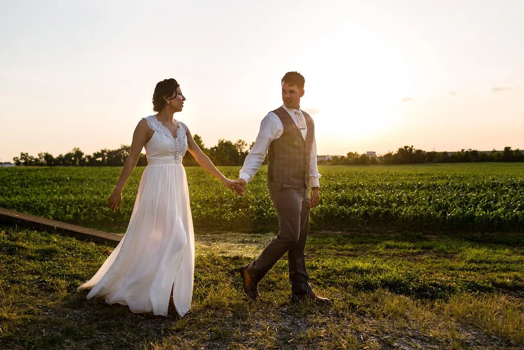 A couple holding hands, walking in a field at sunset, with the woman in a white dress and the man in a vest and dress pants.