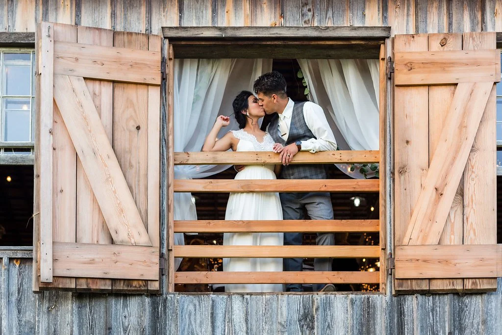 A bride and groom sharing a kiss on a rustic wooden balcony with open shutters.