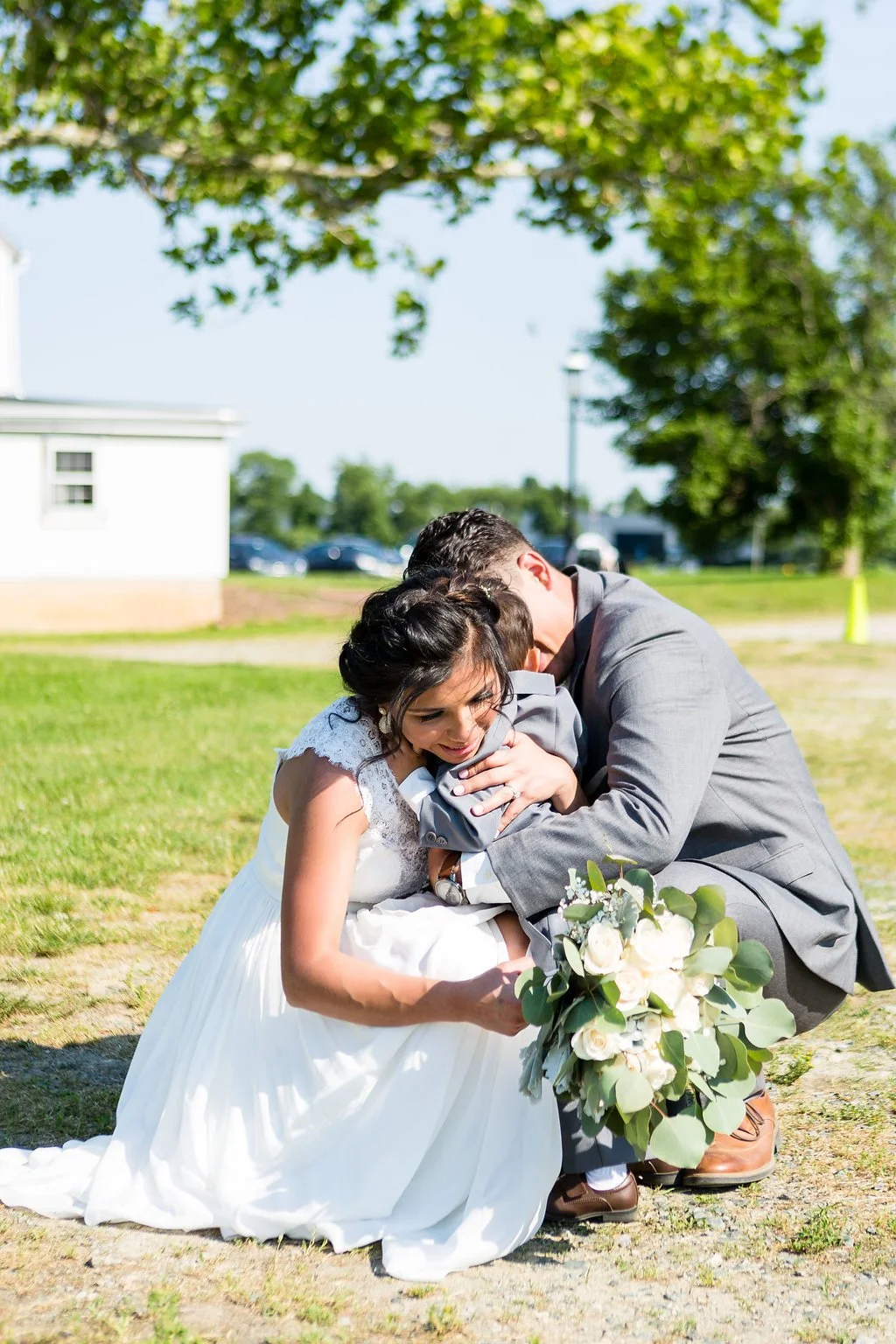 A bride and groom embracing during their wedding outdoors on a sunny day, with the bride holding a bouquet of white roses and greenery.