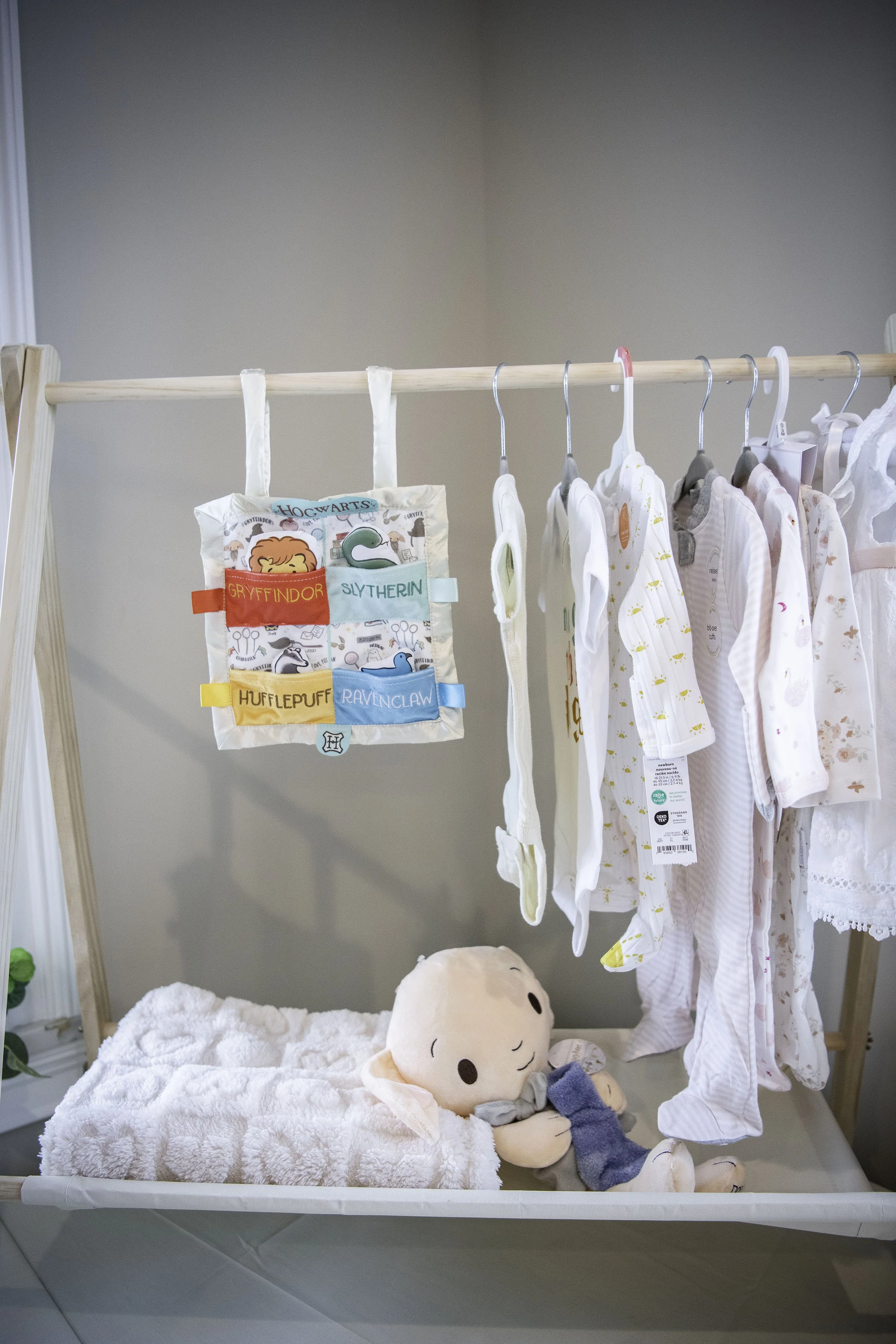Baby clothes hanging on a wooden rack with a plush doll and blanket below.