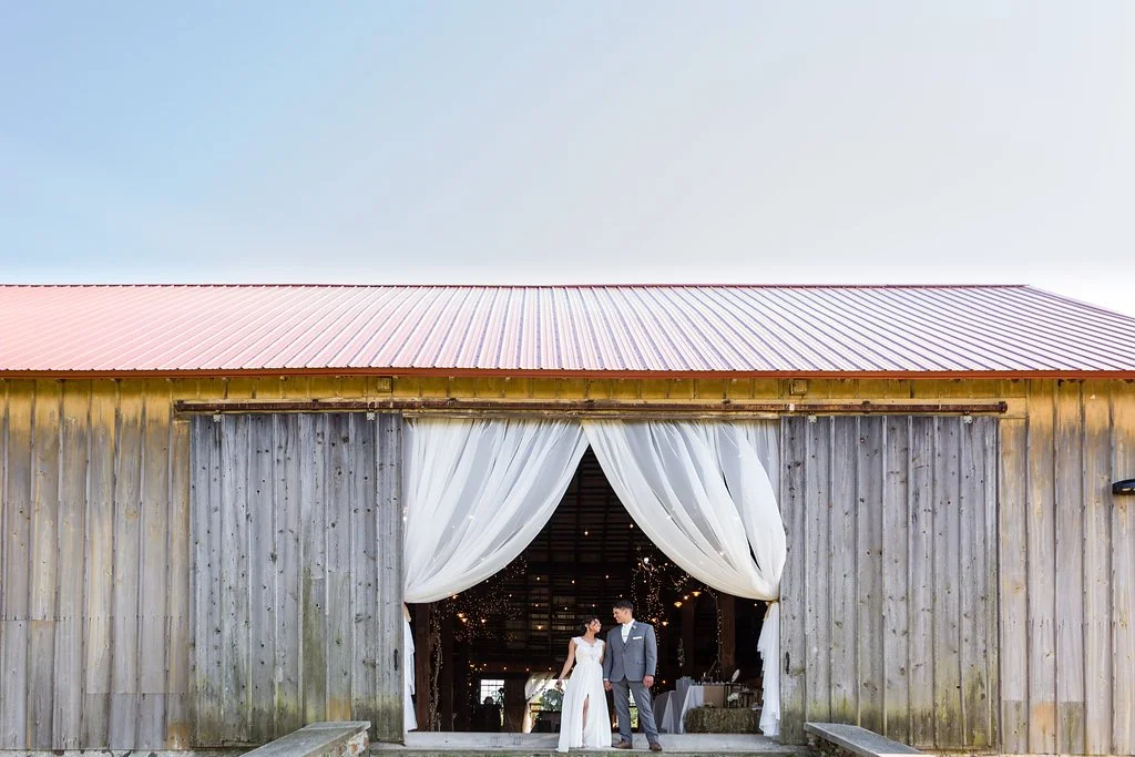 Bride and groom in wedding attire standing in front of a barn with open doors and white curtains.