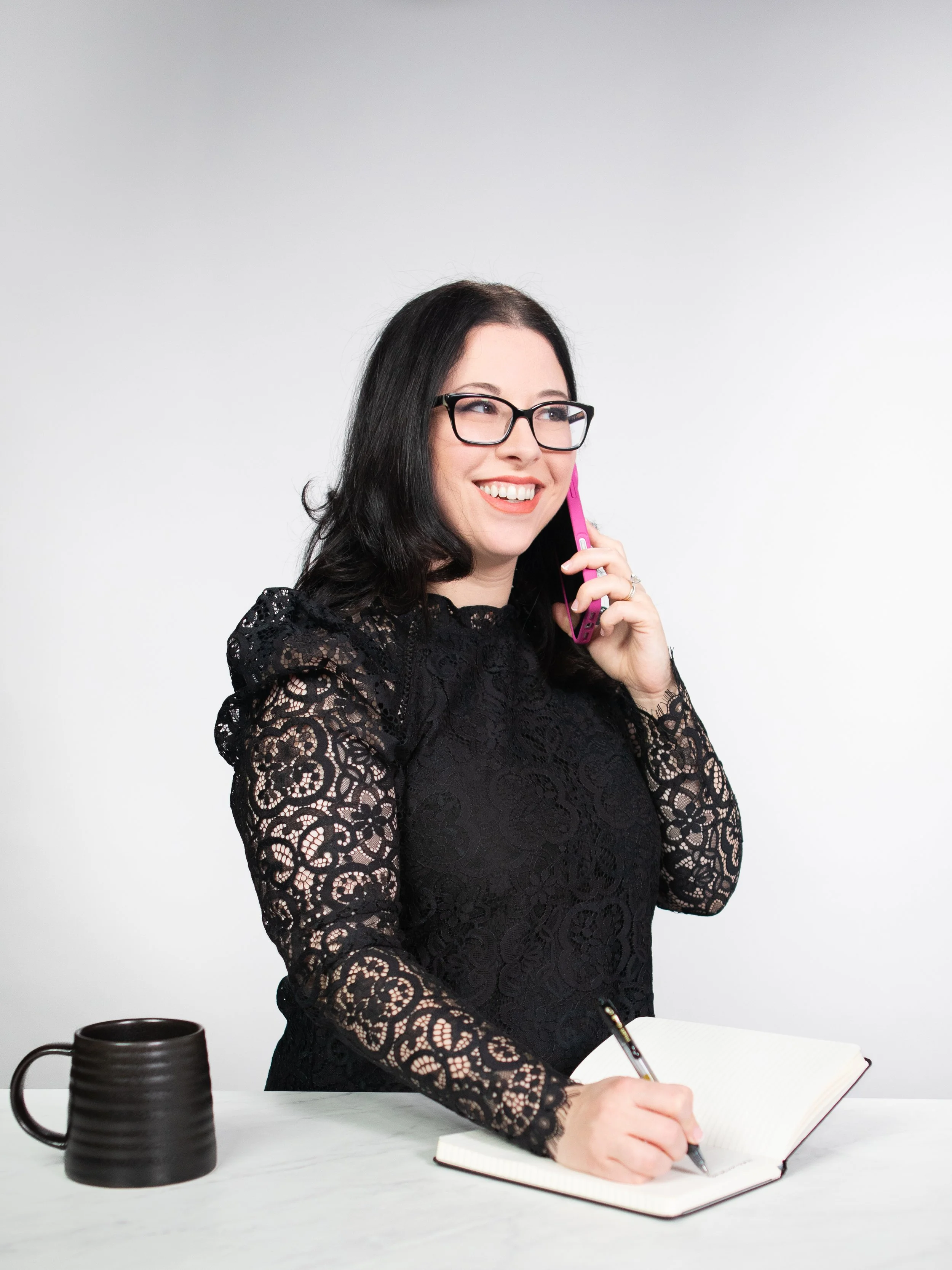 A woman with black hair, glasses, and a black lace dress talking on a pink phone, writing in a notebook, with a black mug on a white table.