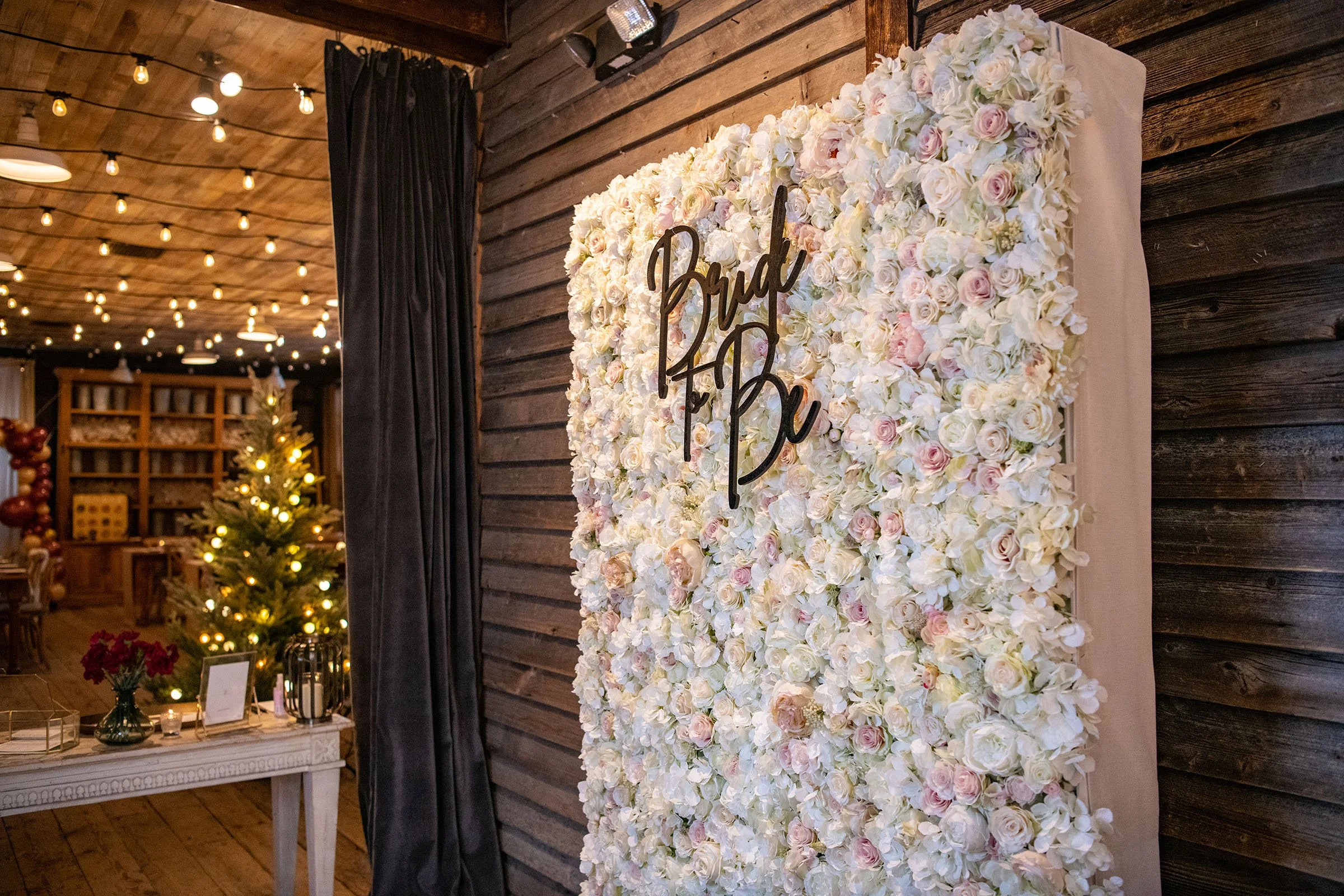 A floral backdrop decorated with white and light pink roses, with a black cursive sign that reads 'Pour & Be'. Behind the backdrop, there is a decorated room with a small Christmas tree with lights, a table with flowers and decorative items, and string lights hanging from the wooden ceiling.