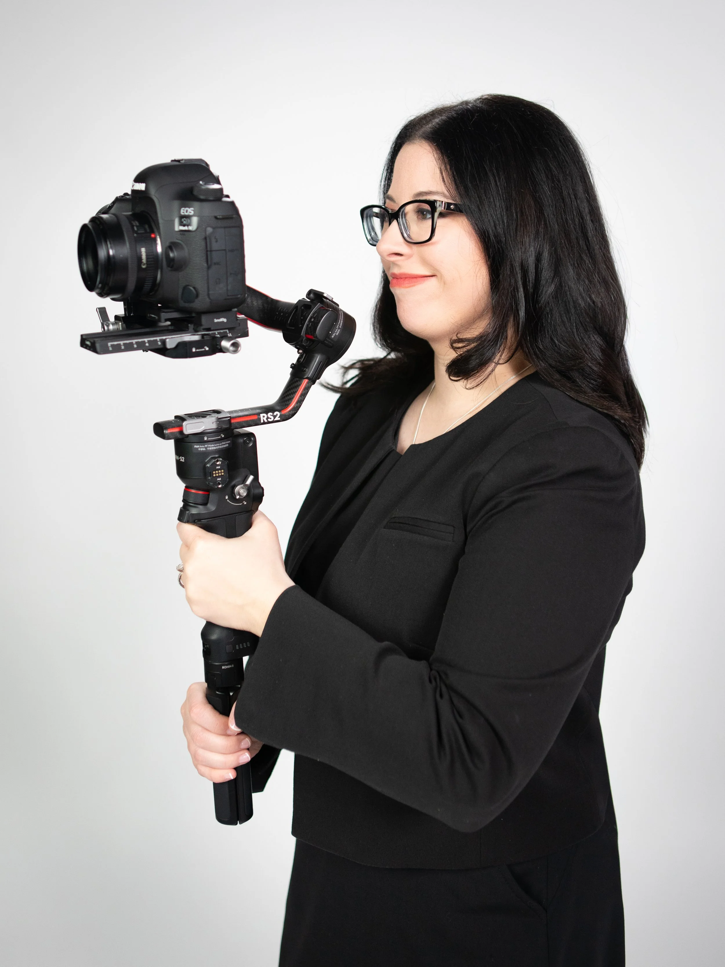 A woman with dark hair and glasses holding a professional camera mounted on a stabilizer against a plain white background.