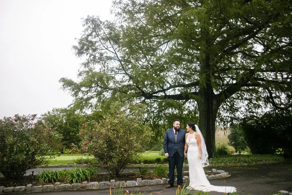 Bride and groom standing together outdoors under a large tree, smiling at each other, in wedding attire.