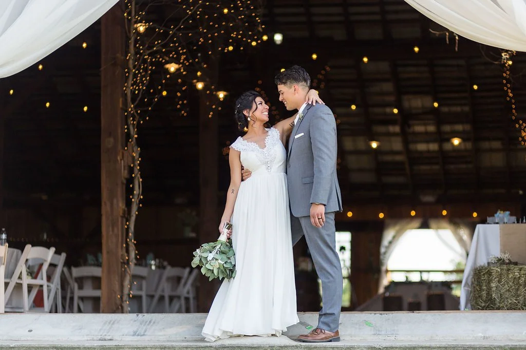 A bride and groom standing close together, smiling at each other during their wedding reception. The bride is holding a bouquet of greenery, wearing a white wedding dress, and the groom is dressed in a gray suit. The setting is a rustic indoor venue 
