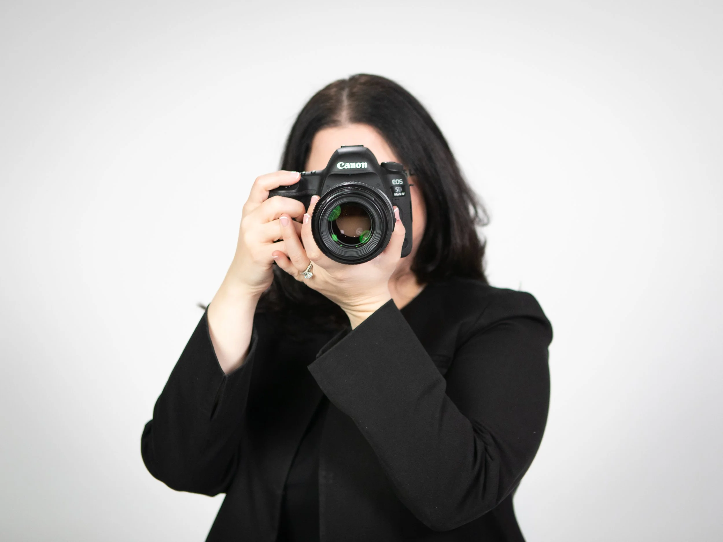 Woman in black blazer taking a photo with a Canon DSLR camera against a plain white background.