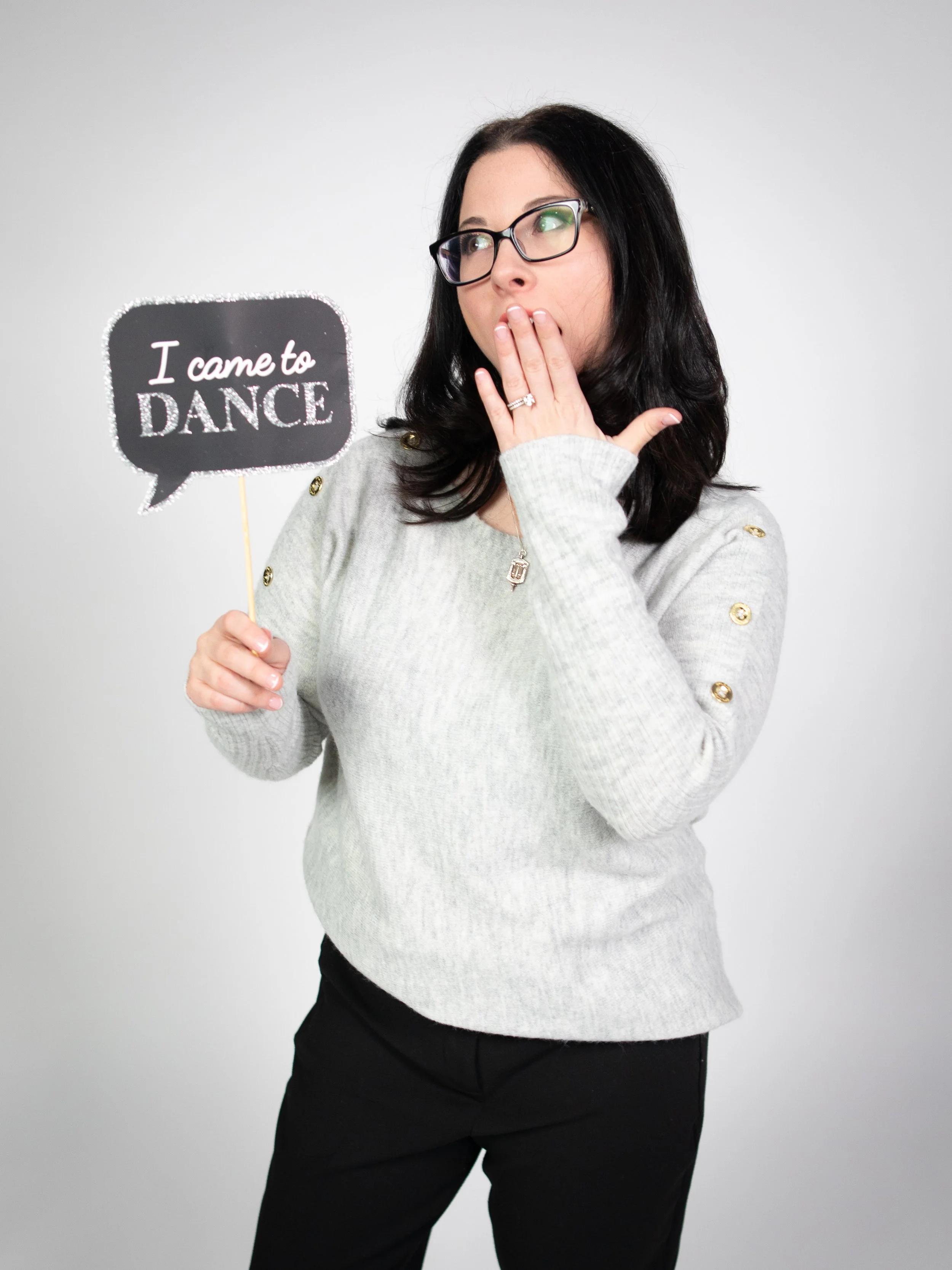 Woman with black hair, glasses, in gray sweater with gold buttons, holding a speech bubble sign that says "I came to DANCE," against a plain gray background.