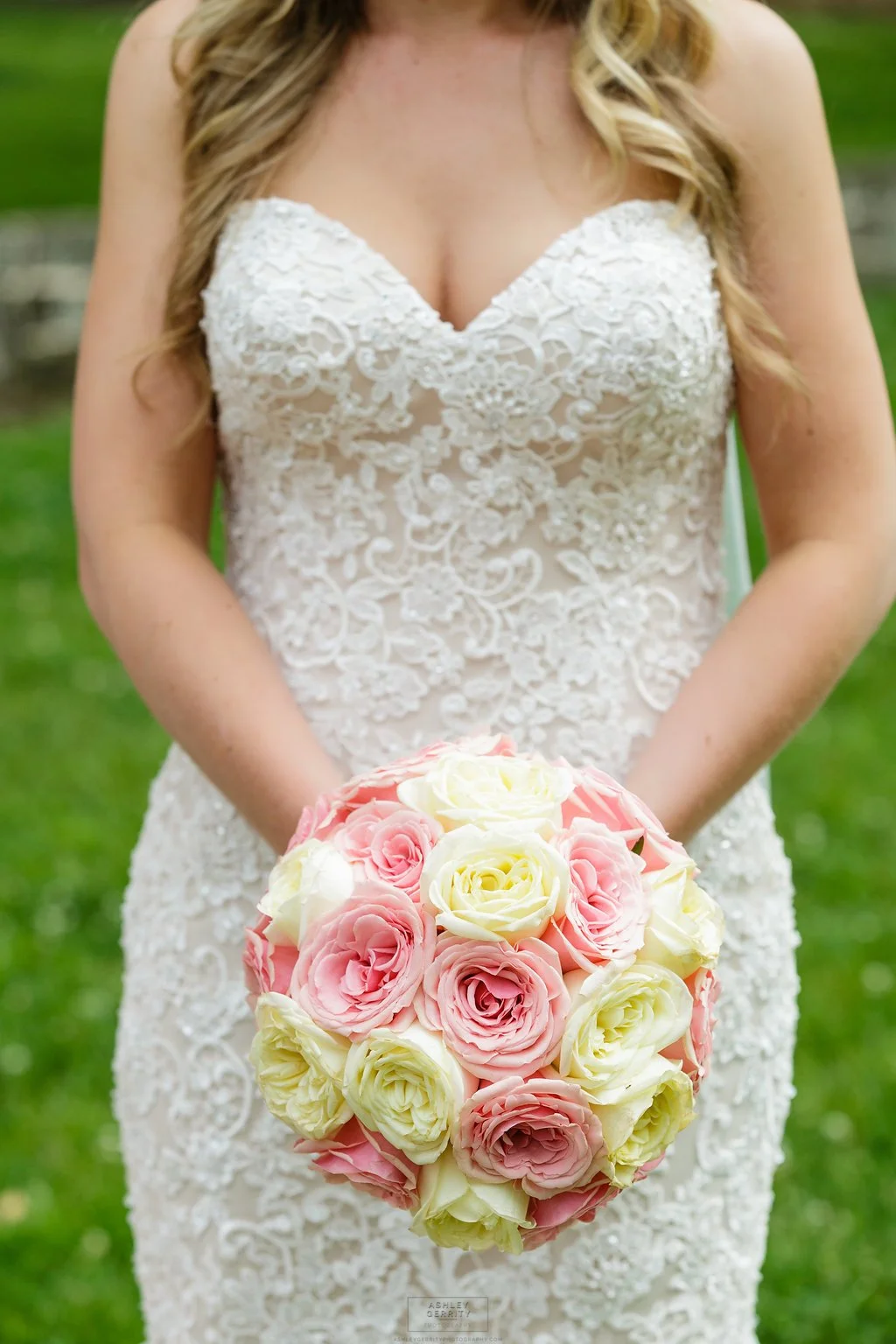 A bride in a white lace wedding dress holding a bouquet of pink and white roses outdoors.