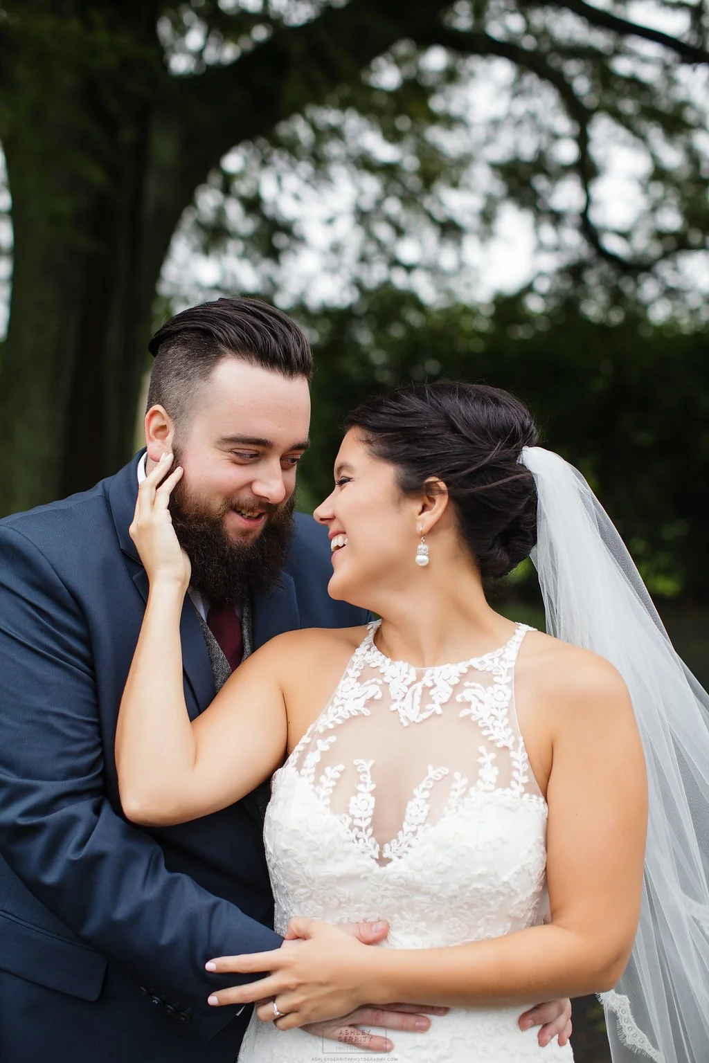 A bride and groom share a joyful moment outdoors on their wedding day, with the groom touching the bride's face and both smiling warmly.