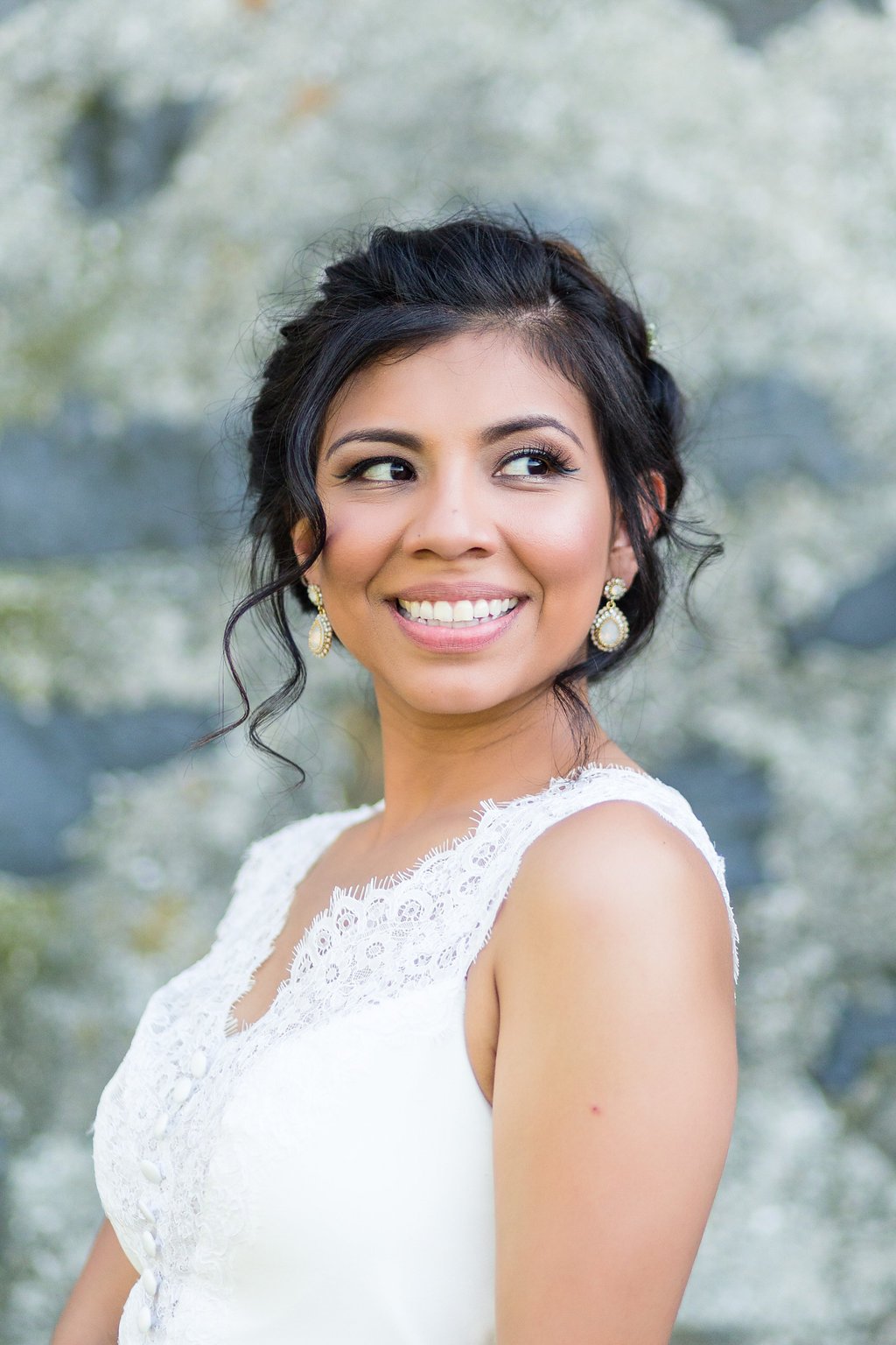 A smiling woman with dark hair styled in loose curls, wearing a white lace dress and chandelier earrings, standing outdoors with a blurred natural background.