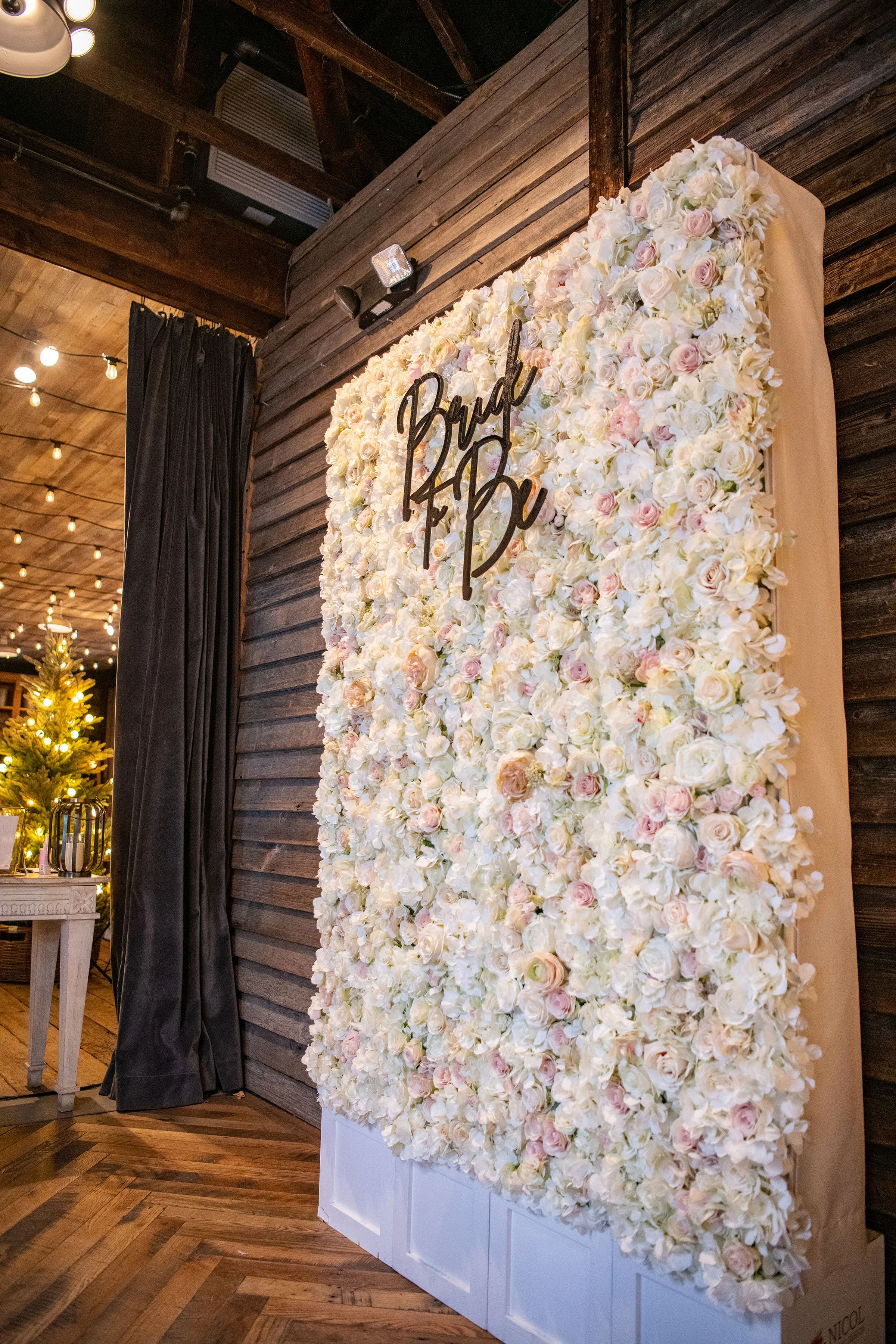 A floral backdrop of white and blush roses with a wooden sign reading 'Bridal Bash' on a beige fabric background, in a rustic indoor venue.